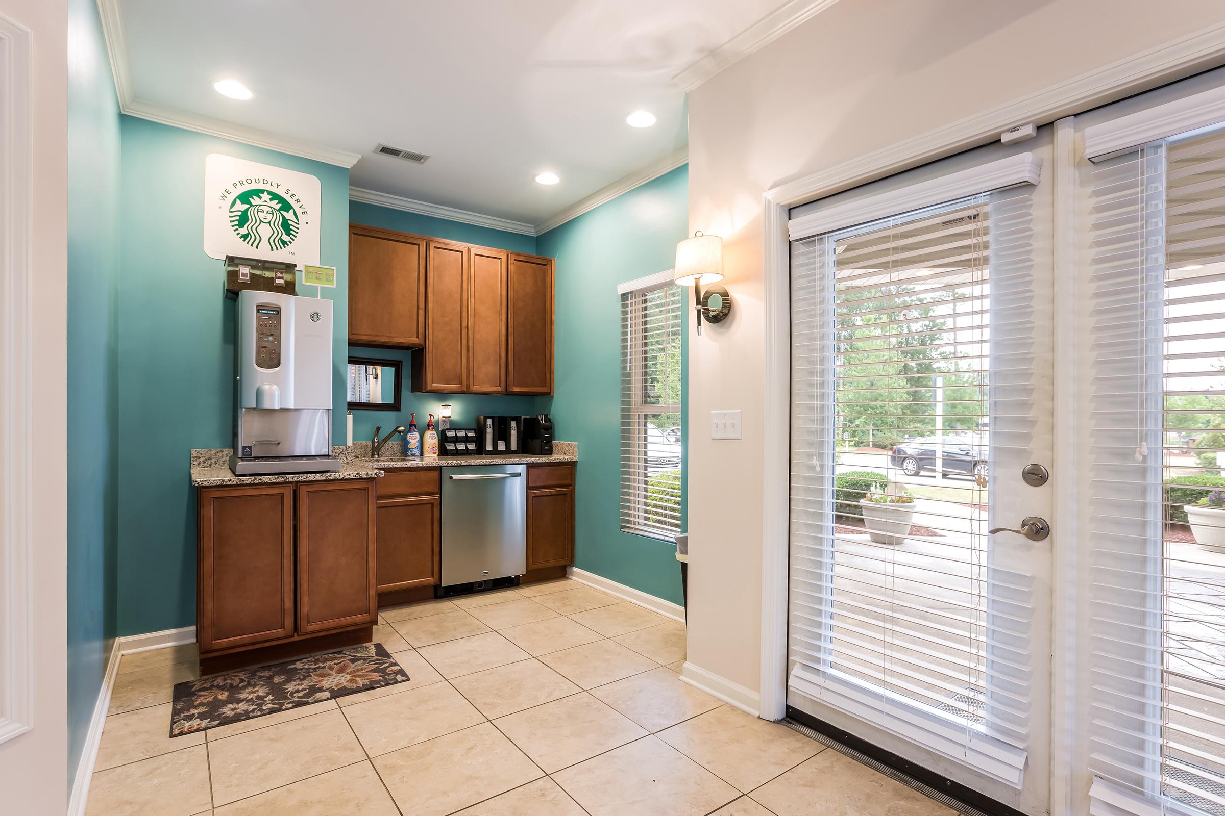 A modern kitchen area featuring wooden cabinets, a granite countertop, and a coffee station with a coffee machine. There are stainless steel appliances, including a dishwasher. A door with blinds leads outside, and a decorative Starbucks sign is on the wall. The floor is tiled, and natural light enters through the door.