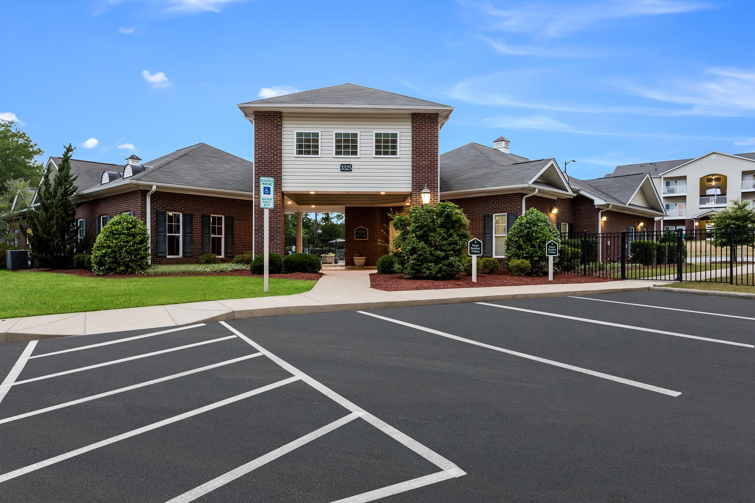 Exterior view of a residential complex entrance, featuring a brick building with a peaked roof, surrounded by well-manicured lawns and shrubs. A parking area is visible in the foreground, with clearly marked white parking spaces and a blue sky in the background.