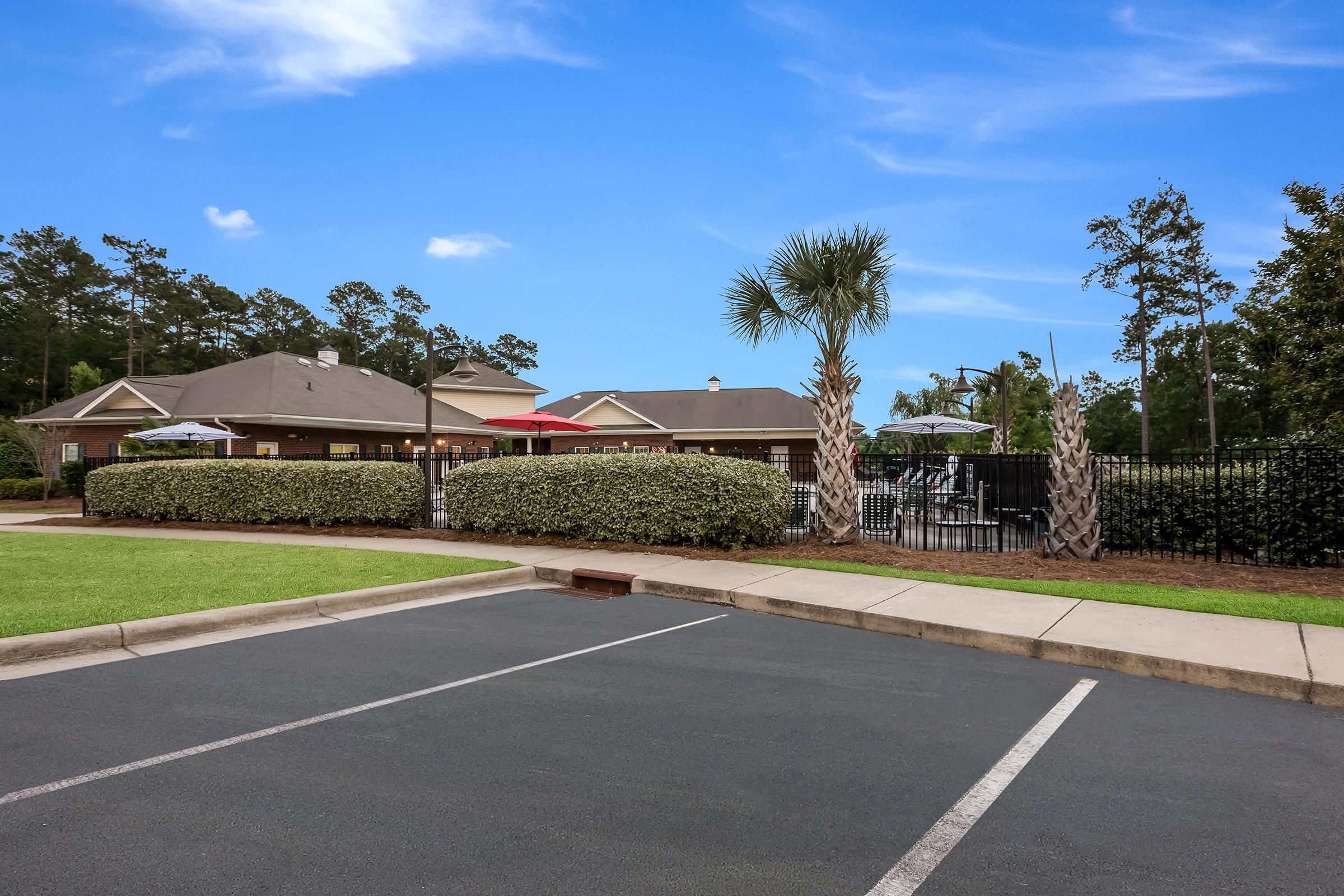 A sunny outdoor area featuring a well-maintained swimming pool surrounded by palm trees and a fence. In the background, there are residential buildings with shaded seating areas. The foreground shows a neatly paved parking lot with empty spaces and green grass on the side, under a bright blue sky.