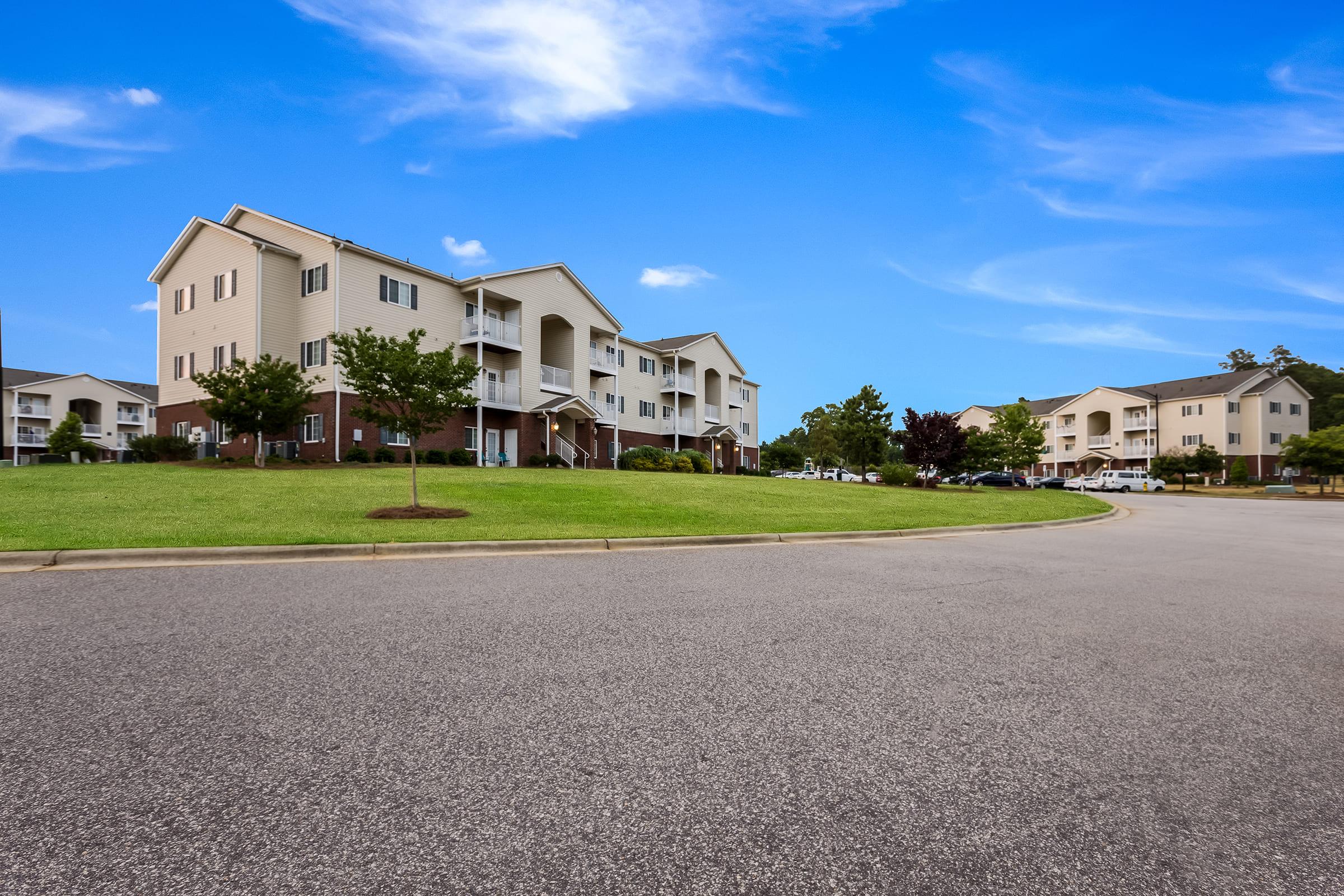 A residential area featuring modern apartment buildings surrounded by green grass and small trees. The clear blue sky complements the scene, while a circular driveway leads to the homes, highlighting a clean, well-maintained community environment.