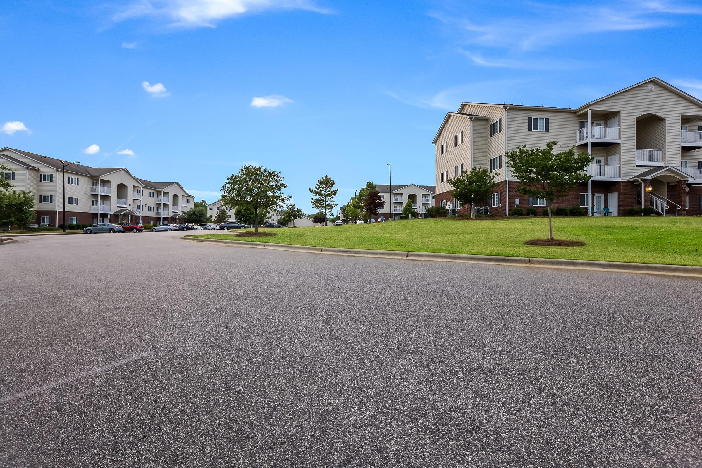 A view of a suburban residential area featuring modern apartment buildings on either side of a wide, empty road. Green lawns and a few trees are visible, under a bright blue sky with scattered clouds. The scene conveys a peaceful and spacious neighborhood atmosphere.