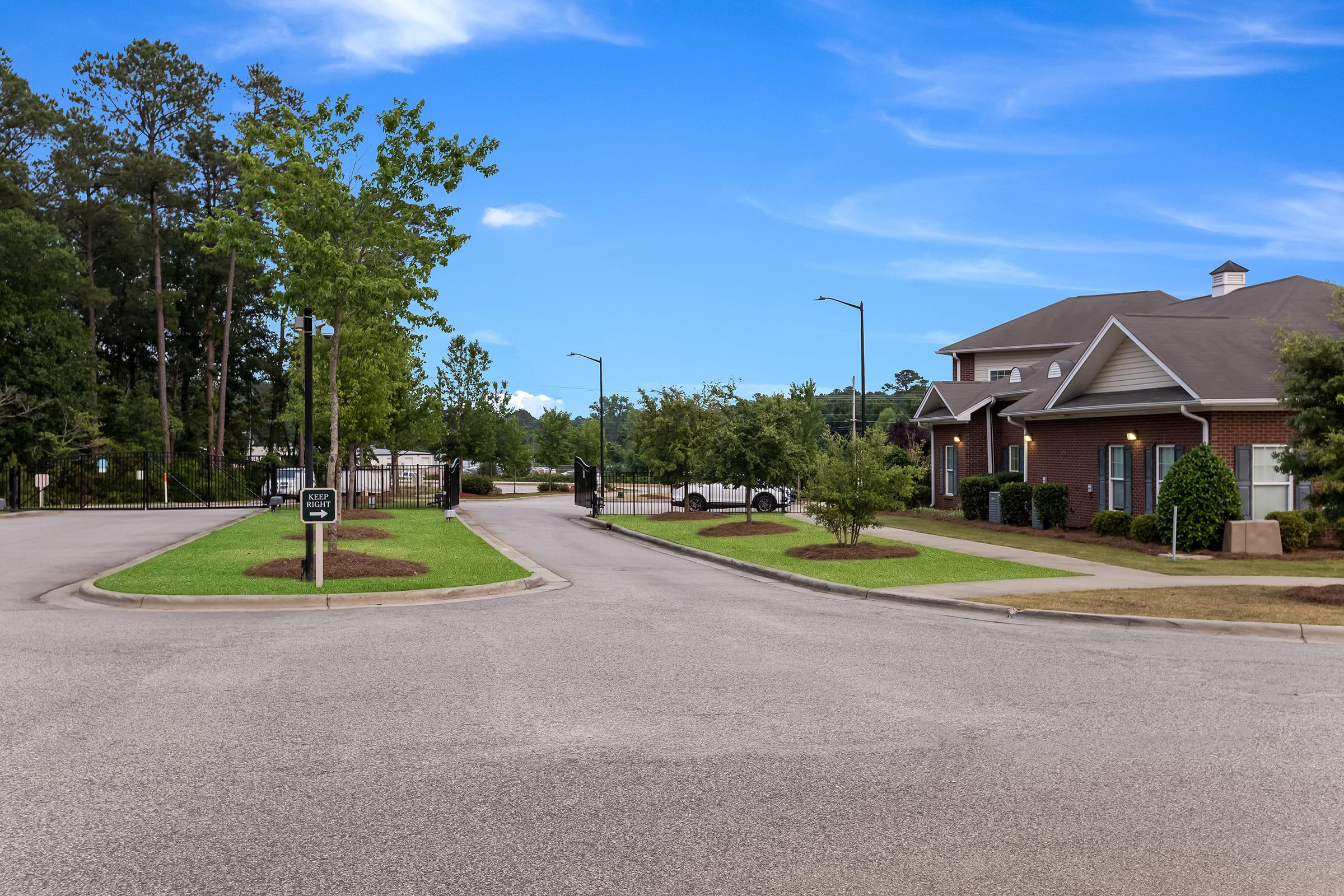 A wide view of a suburban street intersection featuring well-maintained green lawns and trees. To the right, a brick building can be seen, likely a residential or commercial property, with a clear blue sky overhead. Street lamps line the road, and a gated entry is visible in the background.