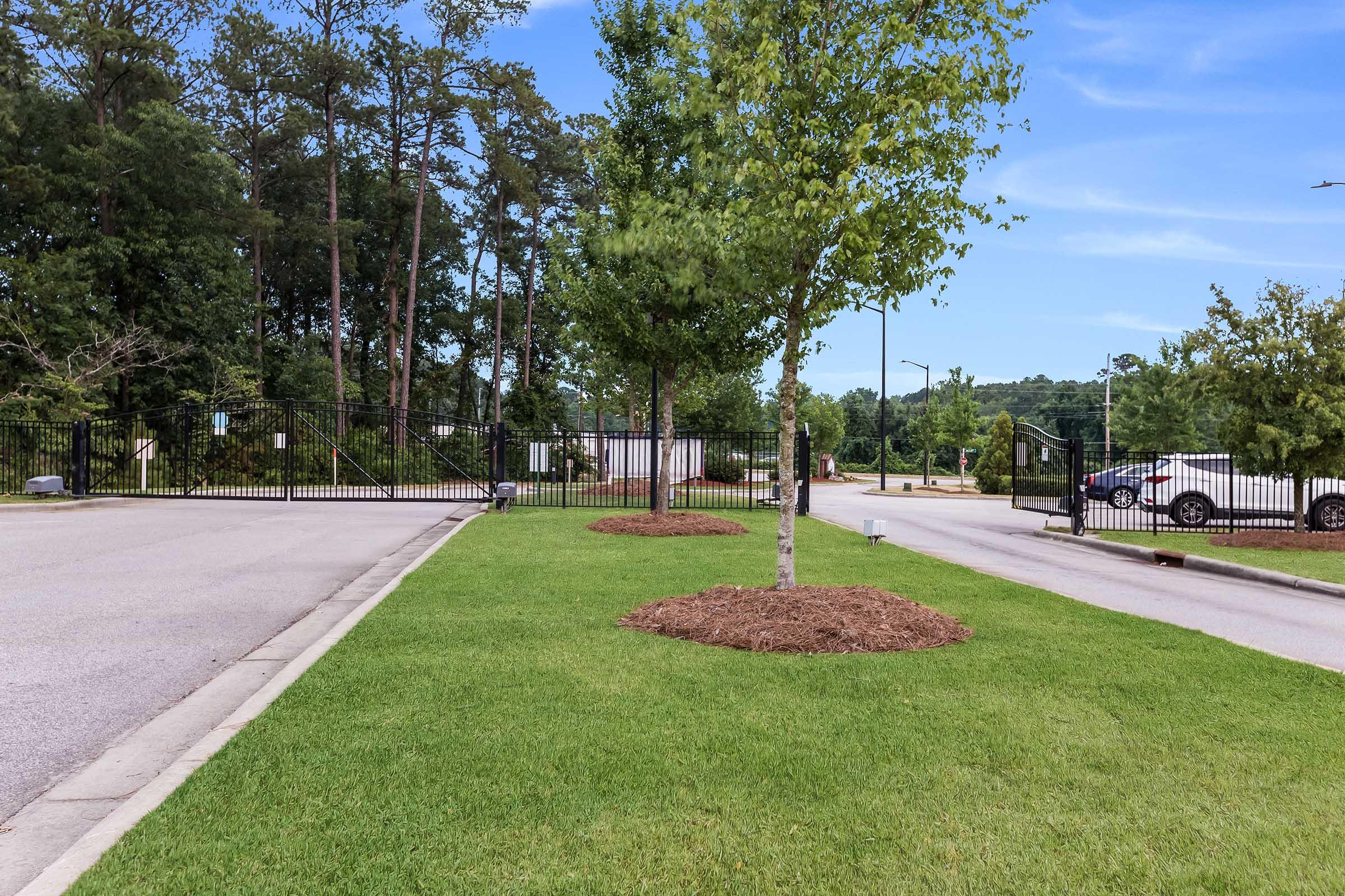 A view of an entrance gate surrounded by greenery, with neatly trimmed grass and young trees. The scene features a two-lane road leading to the gate, flanked by a secure fence and a forested area in the background under a clear blue sky.