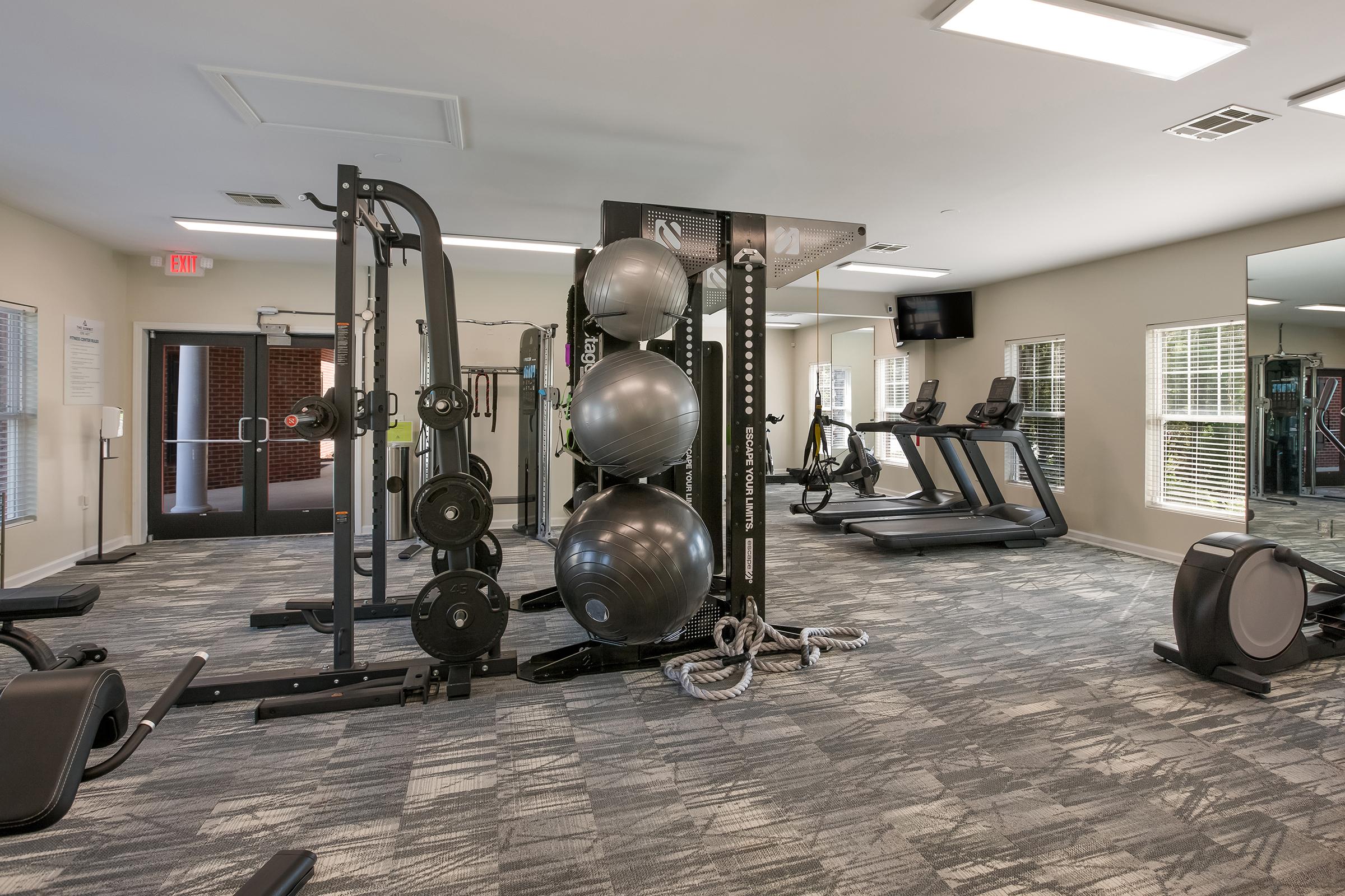 A spacious gym with various exercise equipment, including ellipticals, weight machines, and treadmills. The room features large windows allowing natural light, ceiling fans for ventilation, and multiple wall-mounted televisions. The flooring is black rubber, enhancing the workout environment.