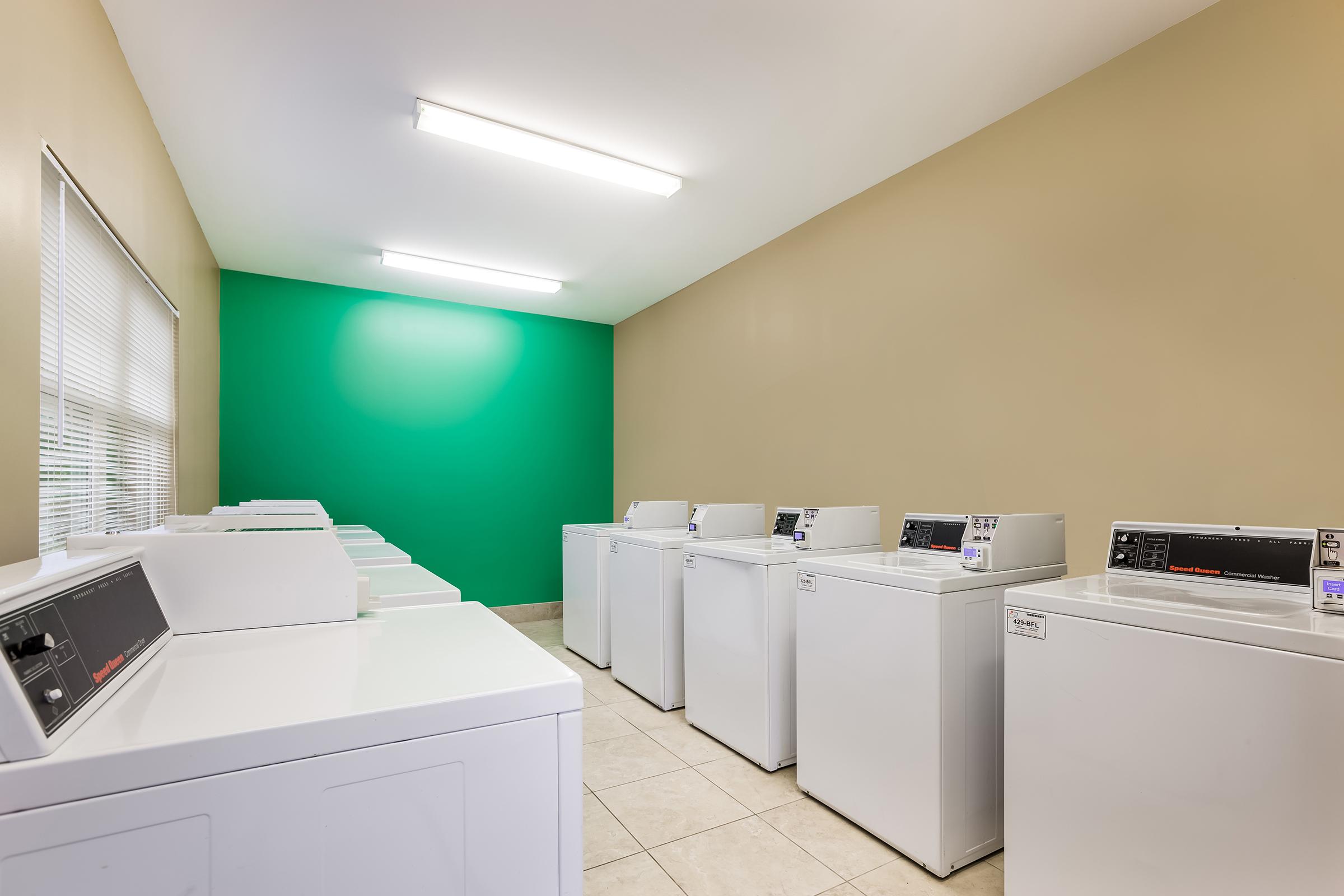 A clean and bright laundry room featuring multiple white washing machines lined up against a beige wall with a vibrant green accent wall. The space is well-lit with overhead fluorescent lighting, and there are large windows providing natural light. The floor is tiled, enhancing the modern appearance.