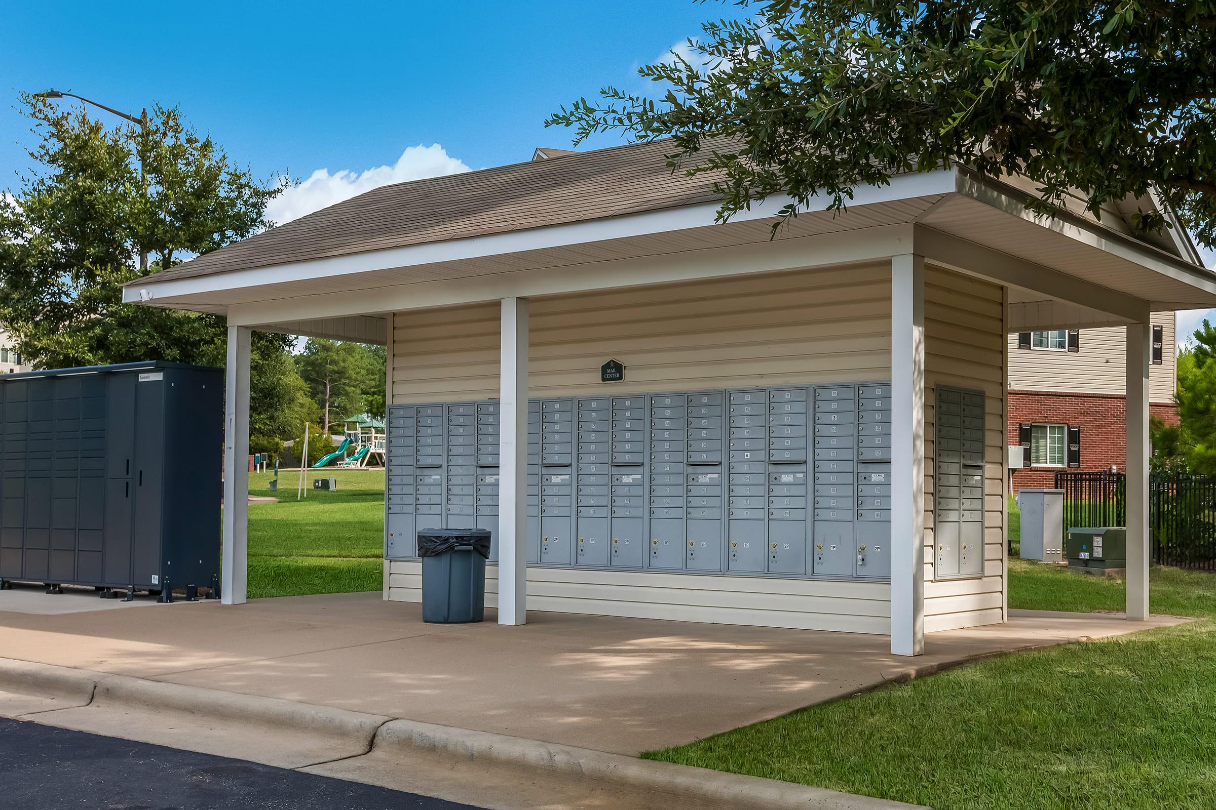 A covered mailbox area featuring multiple locking mailboxes, a large utility storage unit to the left, and a trash can nearby. The structure is situated on a well-maintained lawn with trees and a playground visible in the background, under a clear blue sky.