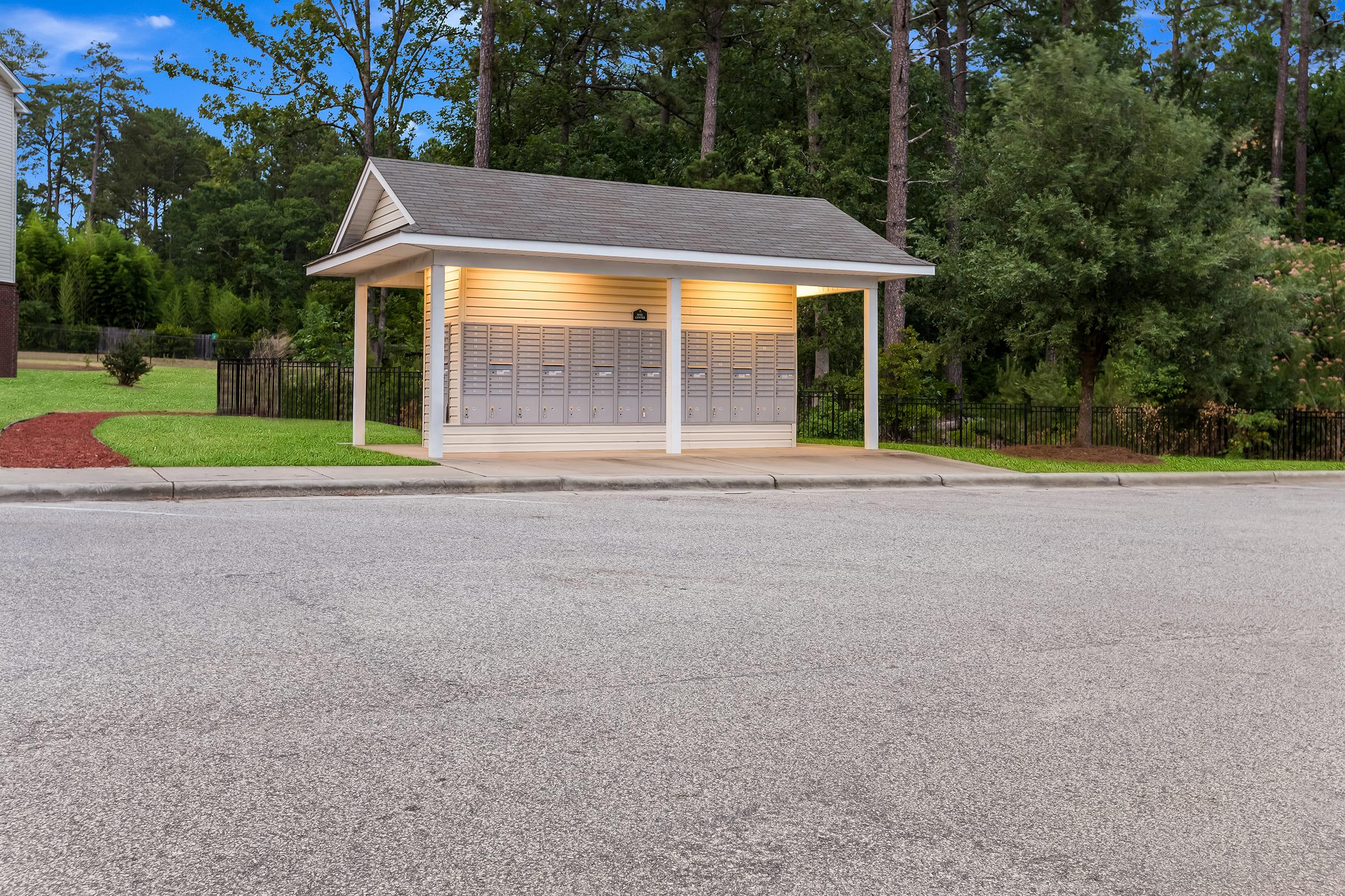 A small, illuminated mail kiosk with multiple mailboxes is situated next to a quiet road. The structure has a sloped roof and is surrounded by greenery and trees, providing a serene setting.