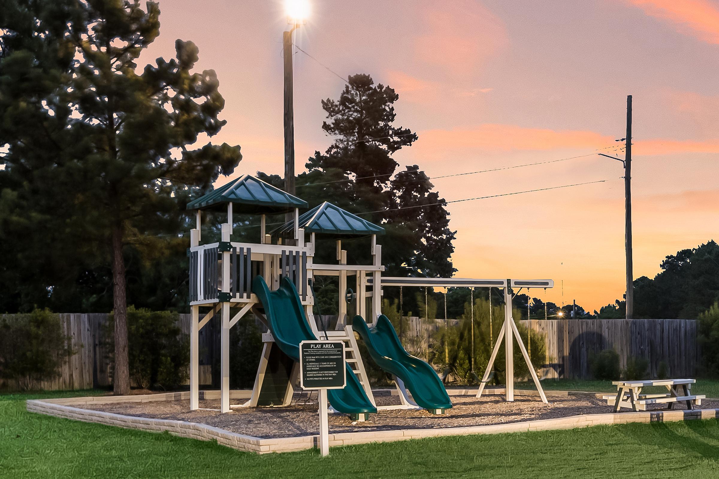 Playground equipment at sunset, featuring two green slides, a swing set, and a picnic table. Surrounded by grass and trees, the scene is illuminated by a nearby light pole, creating a serene atmosphere. A sign indicates playground rules.
