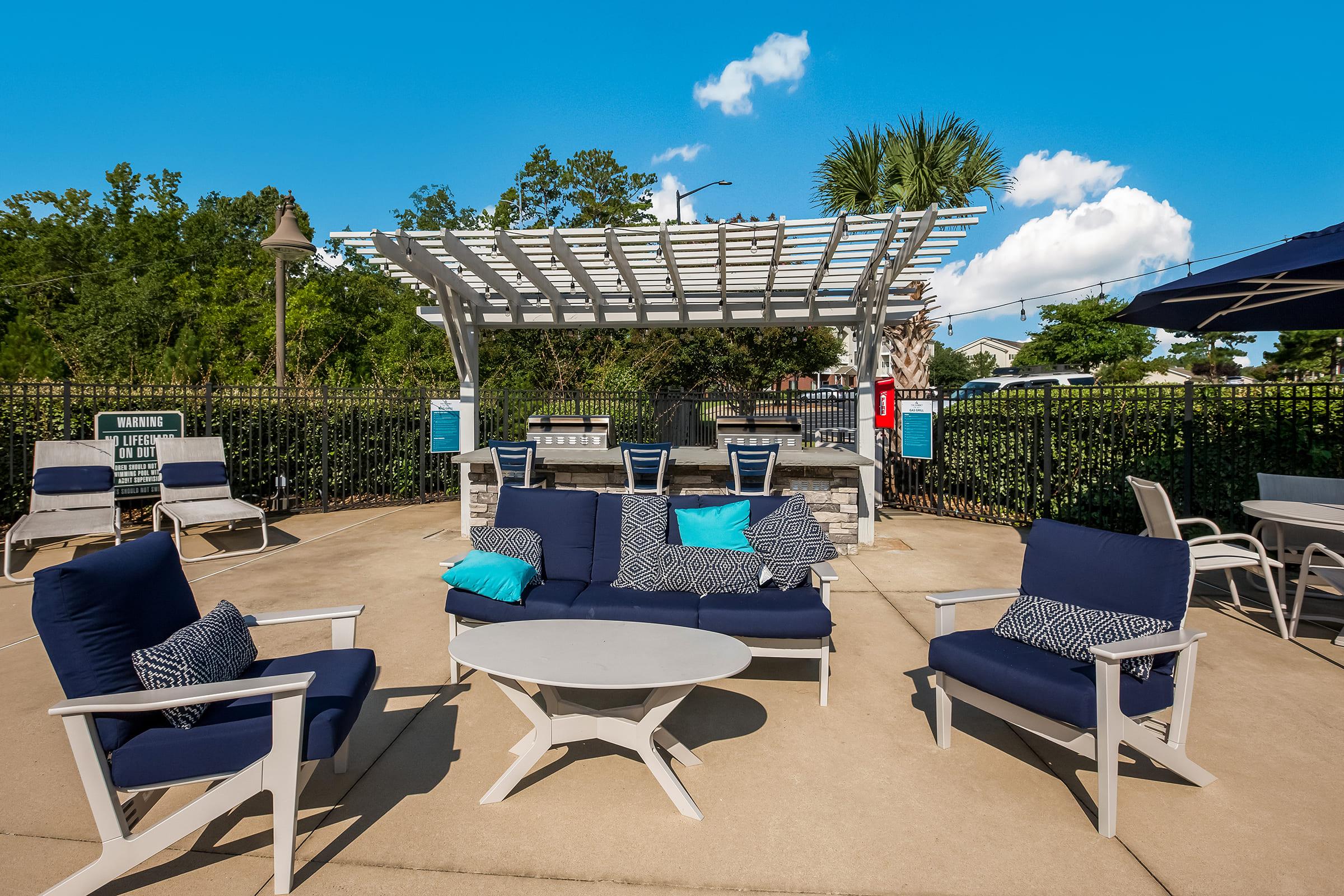 A poolside lounge area featuring navy blue and patterned seating with a round white table. In the background, there are palm trees and a clear blue sky with fluffy clouds. A shaded outdoor cooking space with a grill is visible, surrounded by a well-maintained fence and additional lounge chairs.