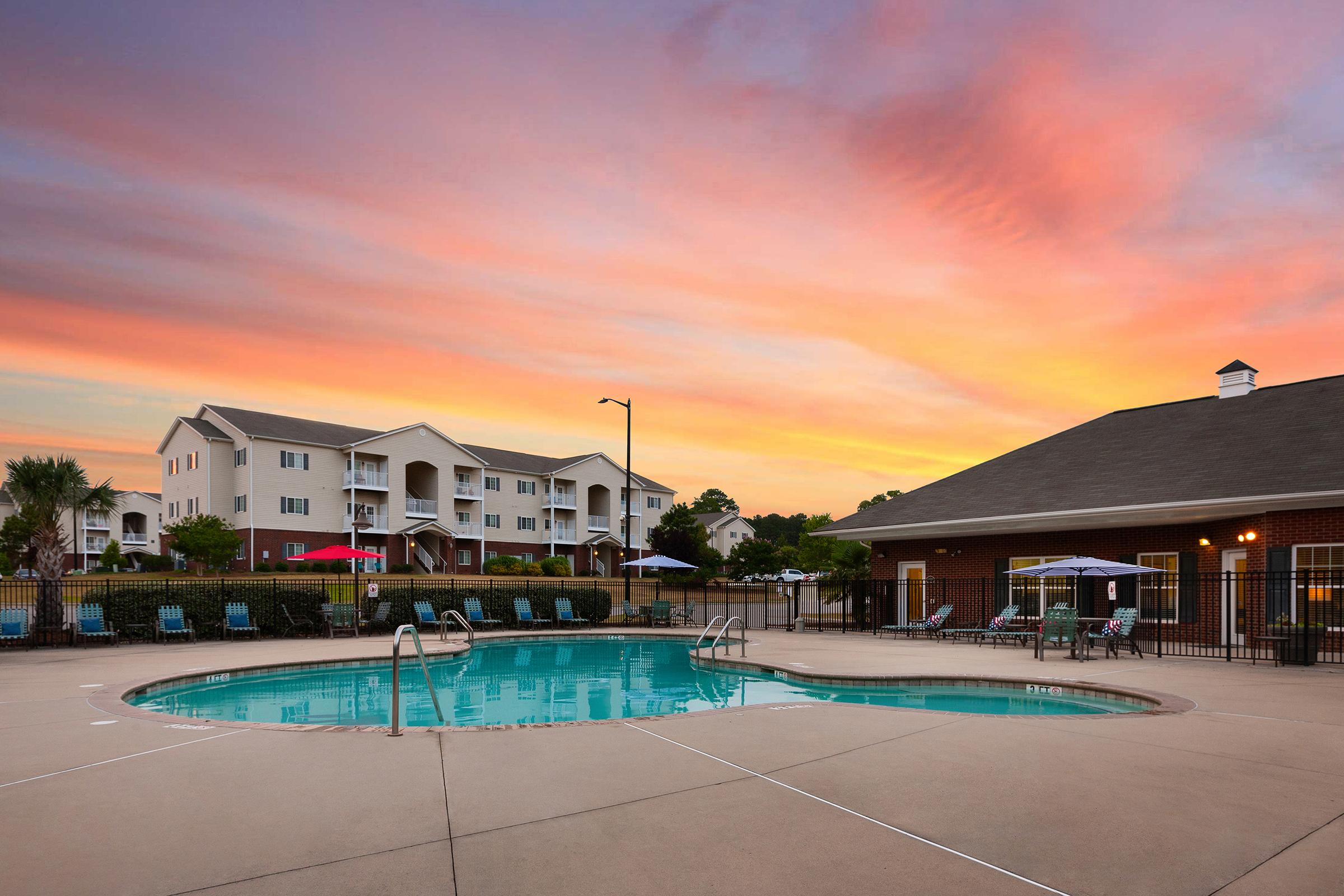 A sparkling swimming pool surrounded by lounge chairs, set against a vibrant sunset sky. In the background, modern apartment buildings create a welcoming atmosphere. The scene conveys relaxation and community, perfect for enjoying warm evenings outdoors.