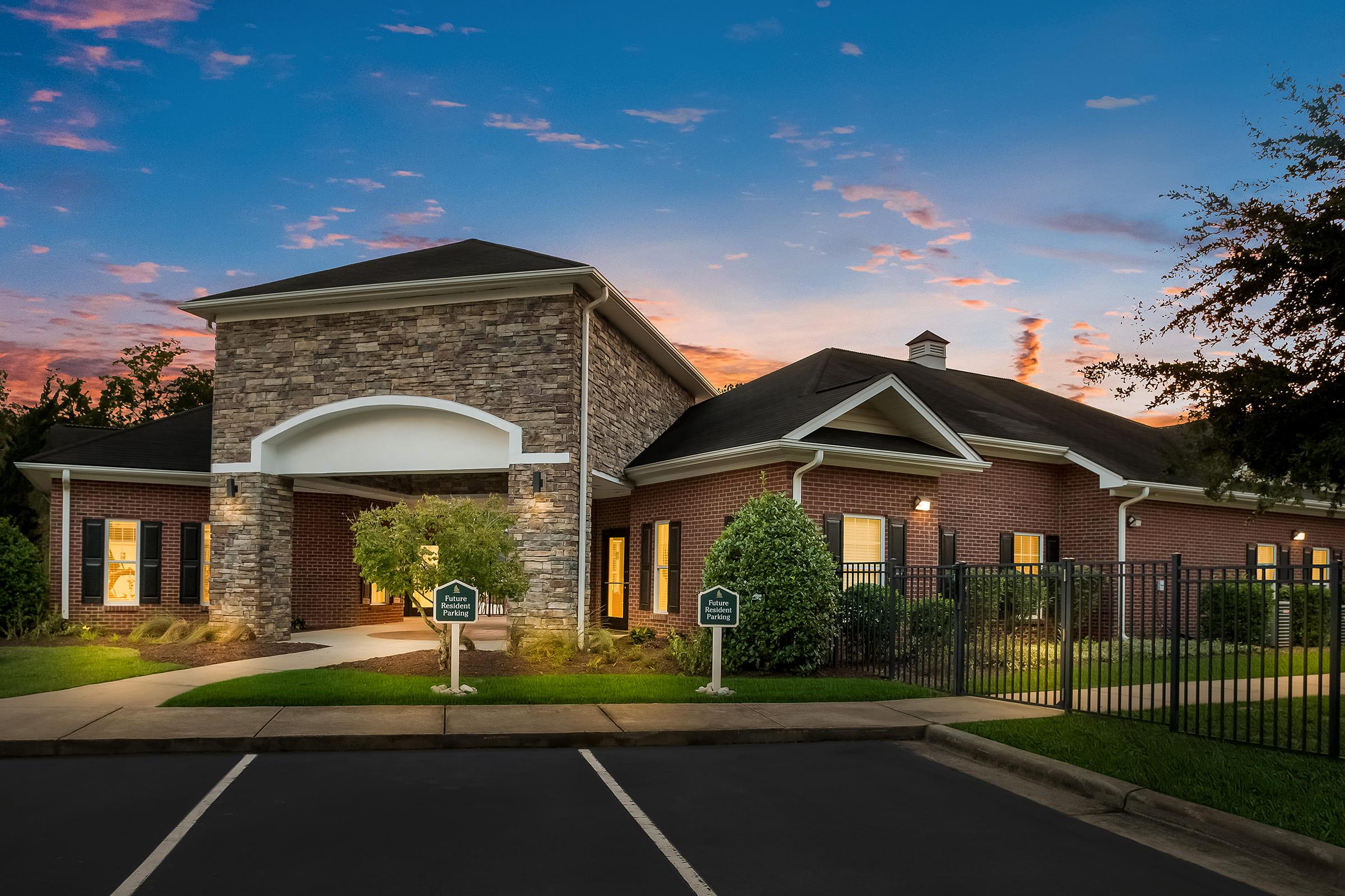 A modern brick building with a stone accent, featuring large windows and well-maintained landscaping. The entrance has a covered awning, and there are signs on either side of the path leading up to the entrance. The sky is a blend of warm colors from a sunset, enhancing the inviting atmosphere of the location.