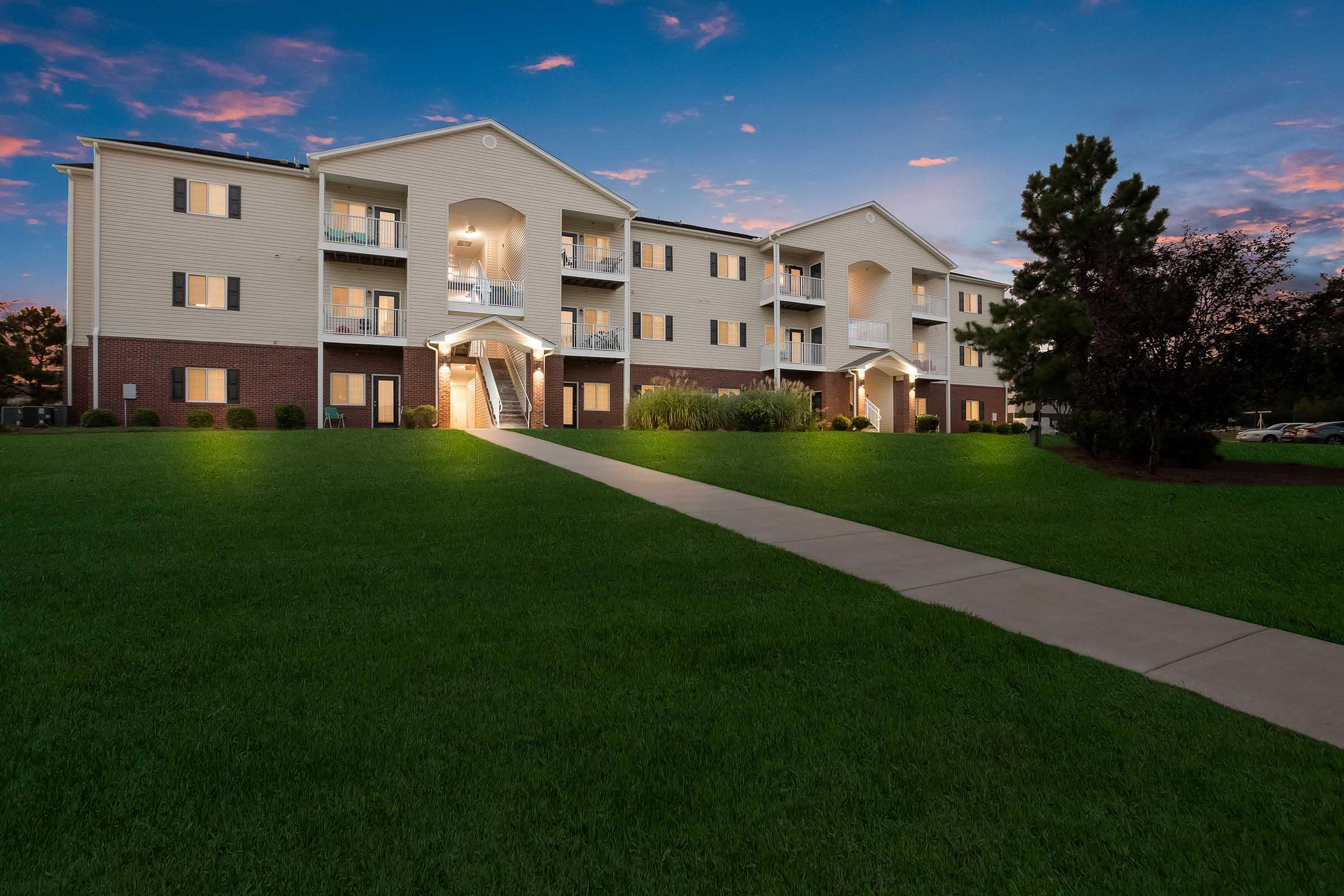 A modern apartment building at twilight, featuring multiple balconies and a well-lit entrance. The surrounding area has lush green grass and a clear sky with soft pink and blue hues, creating a tranquil atmosphere. A paved walkway leads up to the entrance of the building.