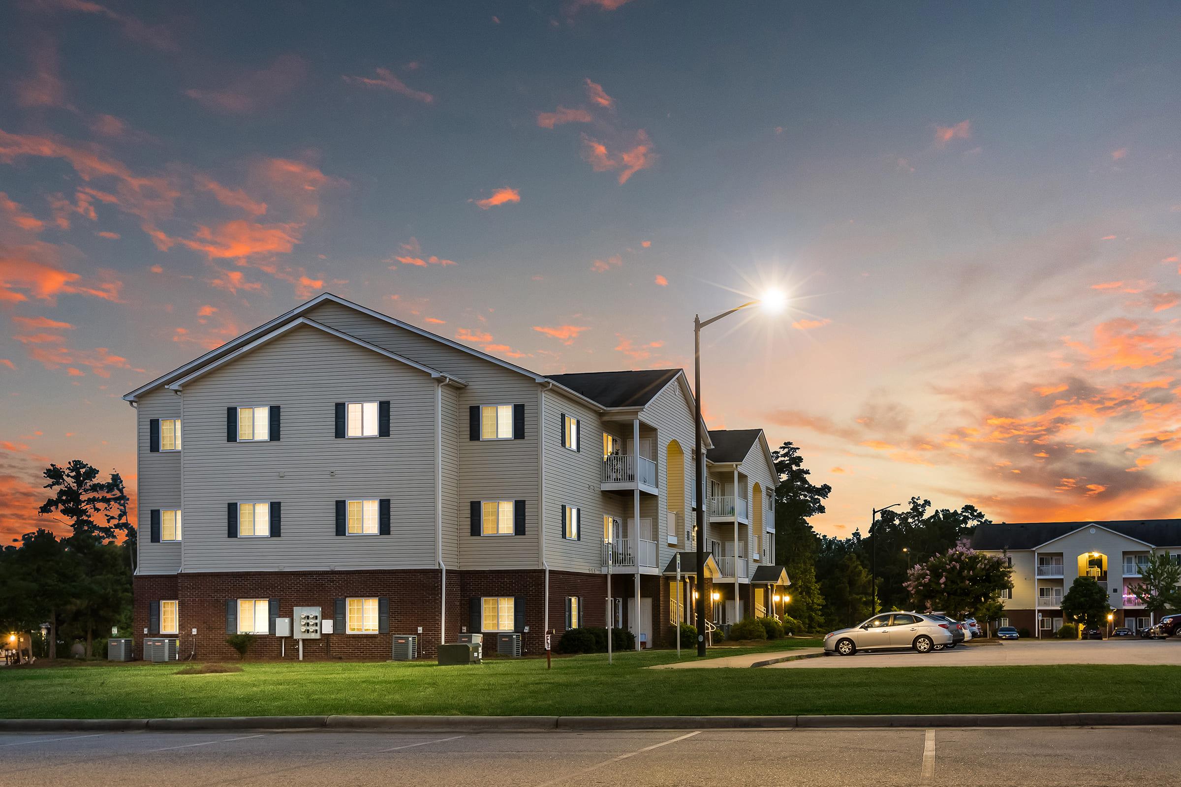 A multi-story residential building at dusk, with several lit windows. The sky is painted in shades of orange and pink, suggesting sunset. Nearby, a white car is parked near the building, with additional trees and another building visible in the background. Streetlights illuminate the area, enhancing the evening atmosphere.