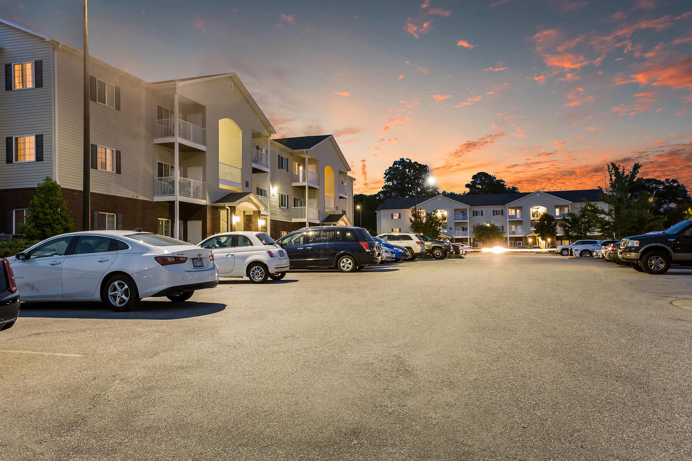 A residential parking lot at dusk, featuring several parked cars and two apartment buildings. The sky is colorful with shades of orange and blue as the sun sets, creating a serene atmosphere. The scene captures a quiet evening in a suburban neighborhood.