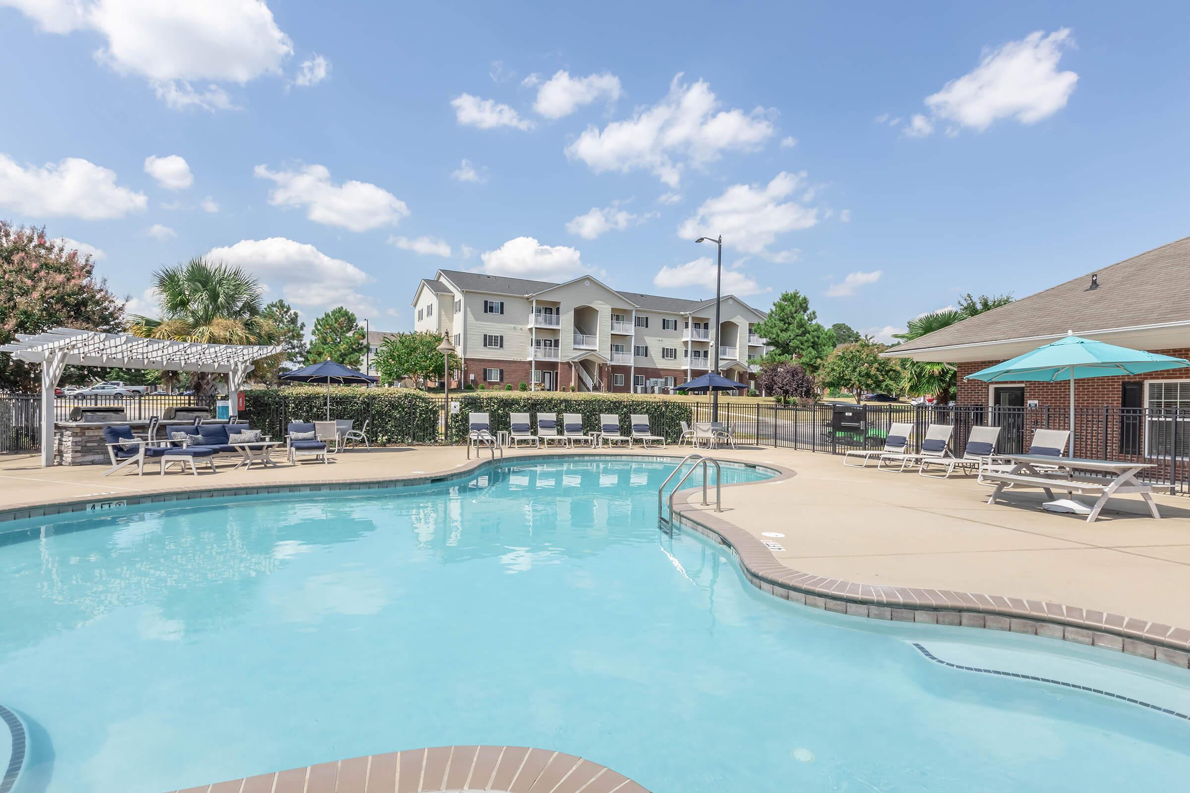 Swimming pool area with clear blue water, surrounded by lounge chairs and umbrellas. In the background, there are residential buildings and greenery under a bright blue sky with fluffy white clouds. A covered seating area with white lattice can be seen near the pool.