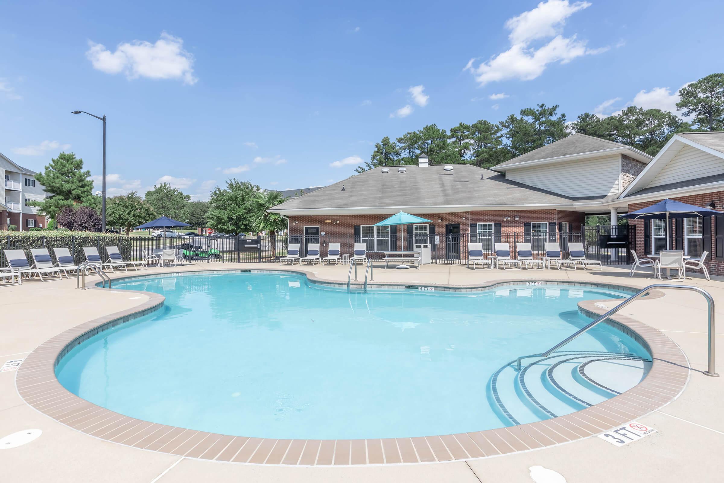 A sparkling blue swimming pool with a gentle slope and surrounding deck chairs under clear blue skies. In the background, there are several trees and an overhanging building, creating a relaxing atmosphere ideal for leisure and recreation.
