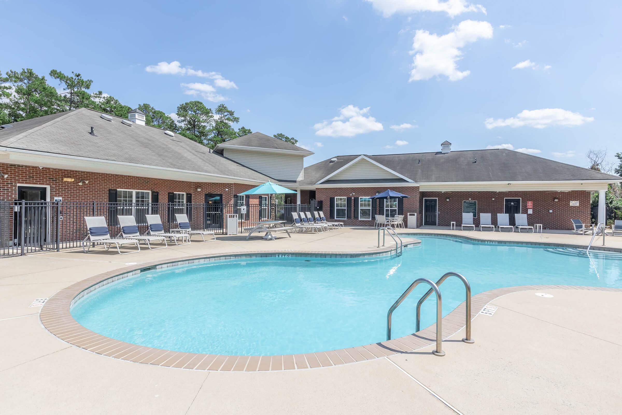 A clear blue swimming pool surrounded by lounge chairs and a patio area. In the background, there is a brick building with multiple windows and a covered area. The sky is bright with a few clouds, and trees are visible in the background, creating a serene environment.