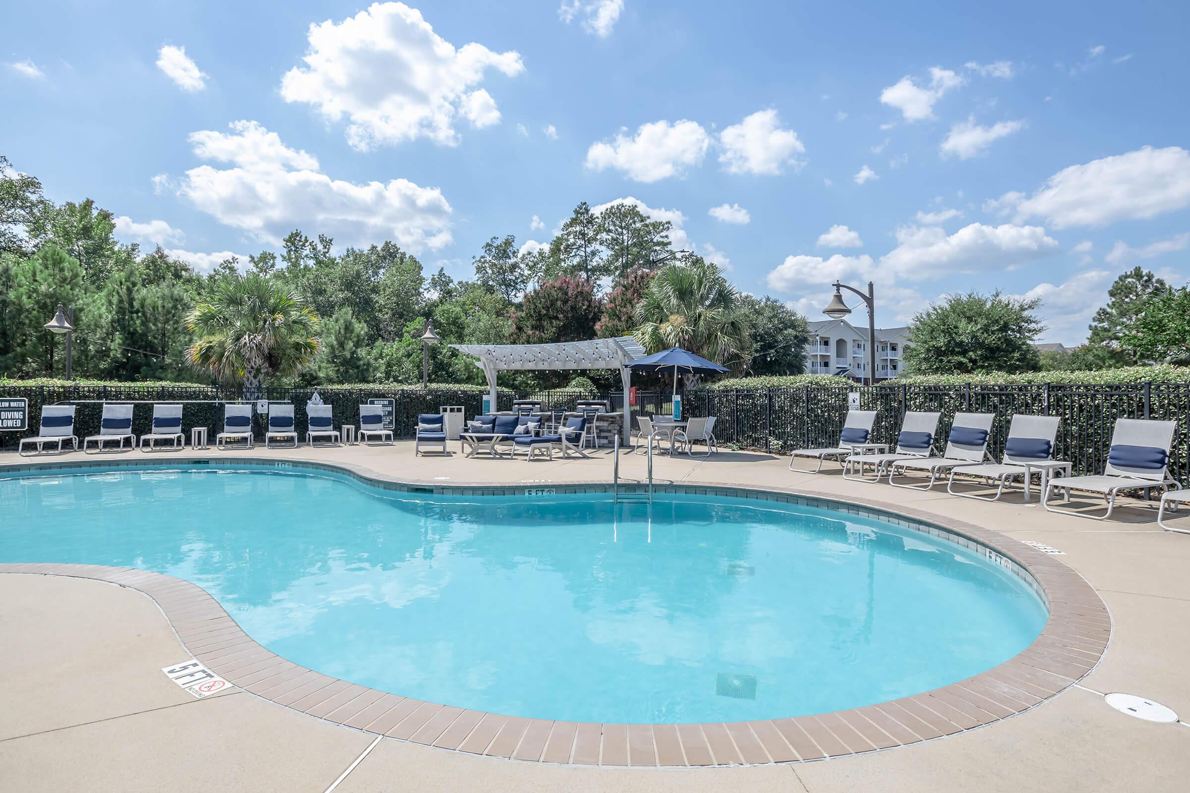 A clear swimming pool surrounded by lounge chairs with a hedge of greenery and trees in the background. The sky is bright blue with fluffy white clouds, and there are umbrellas providing shade near the pool area.