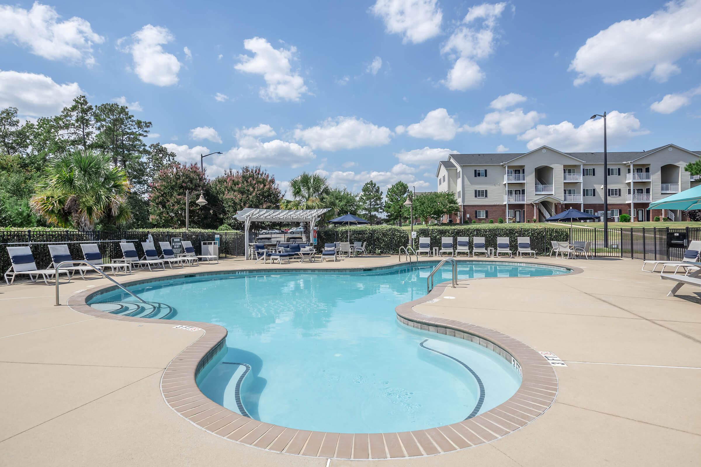 A clear blue swimming pool with a curved edge, surrounded by lounge chairs and a shaded seating area. Lush greenery and a residential building can be seen in the background under a partly cloudy sky.