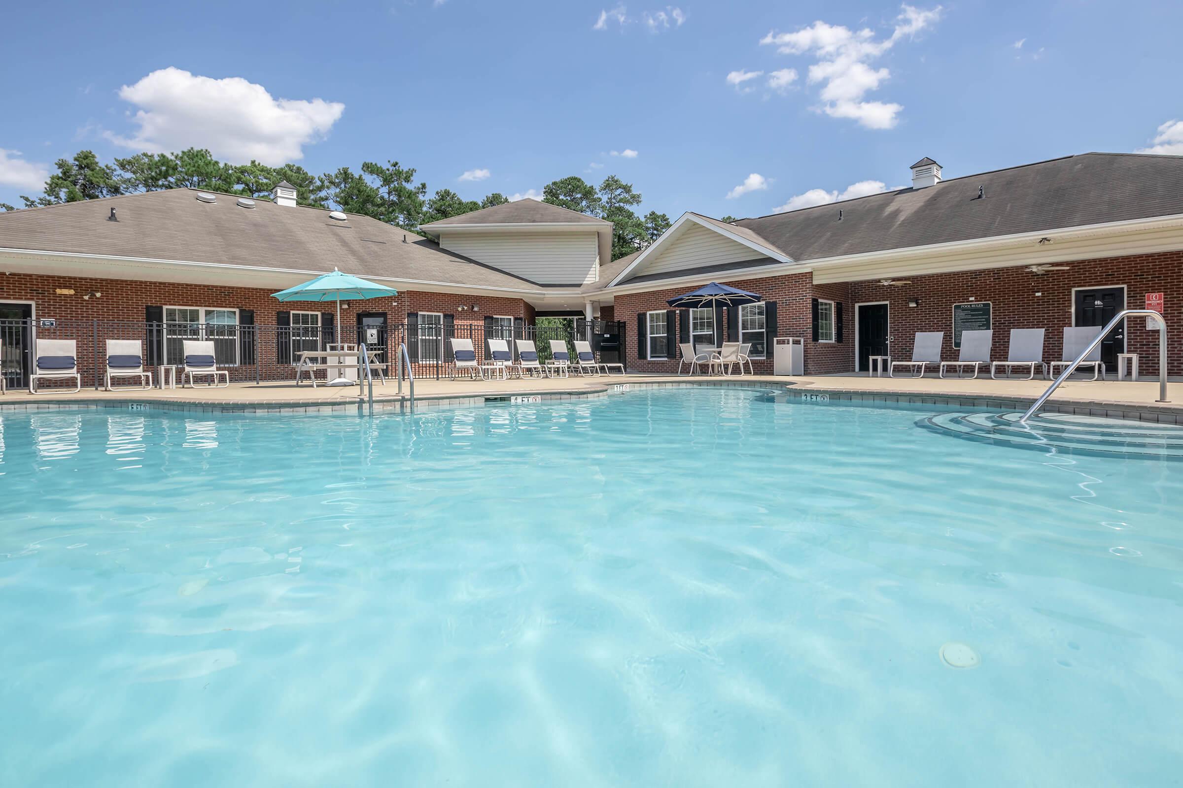 A clear swimming pool with a light blue surface, surrounded by lounge chairs and umbrellas. In the background, there are brick buildings with large windows, under a bright blue sky with scattered clouds. The scene conveys a relaxed, inviting atmosphere suitable for leisure and enjoyment.