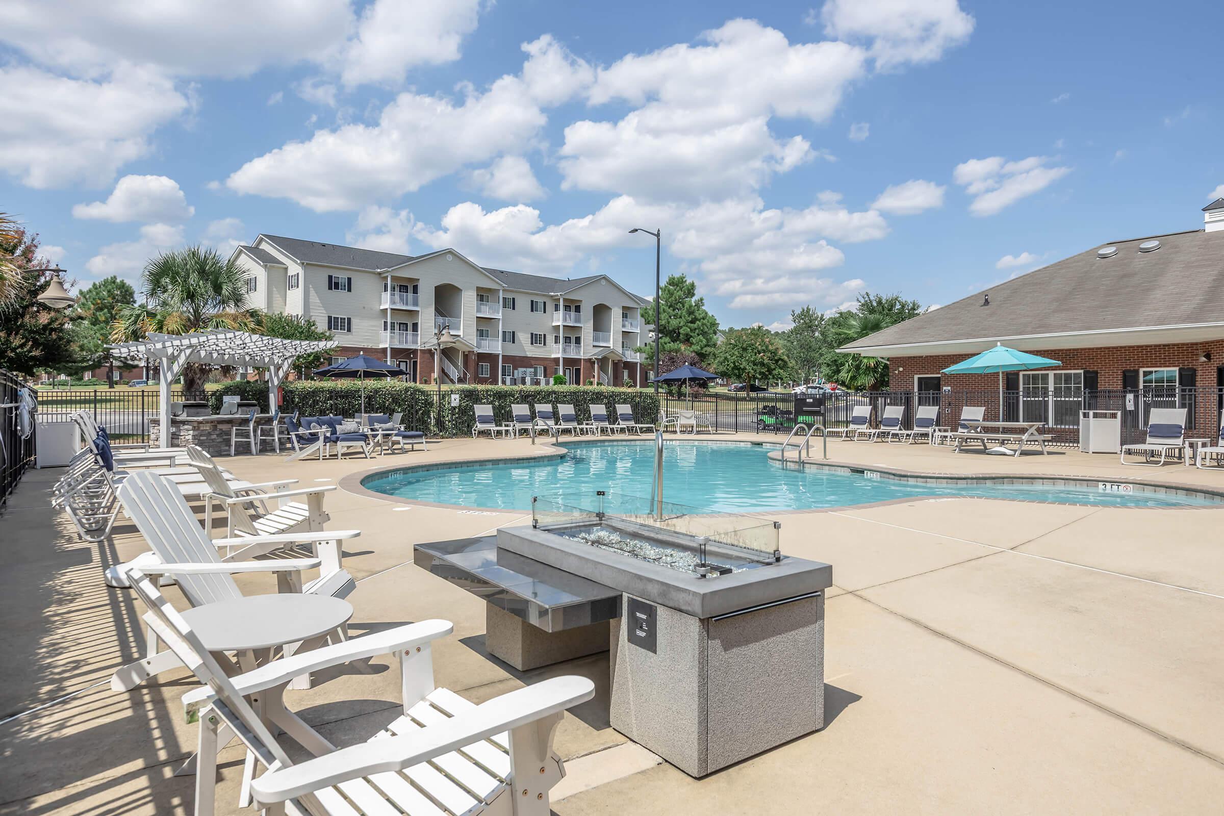 A spacious outdoor swimming pool with lounge chairs and a grill area, surrounded by trees and residential buildings. The sky is blue with fluffy clouds, creating a bright and inviting atmosphere for relaxation and leisure.