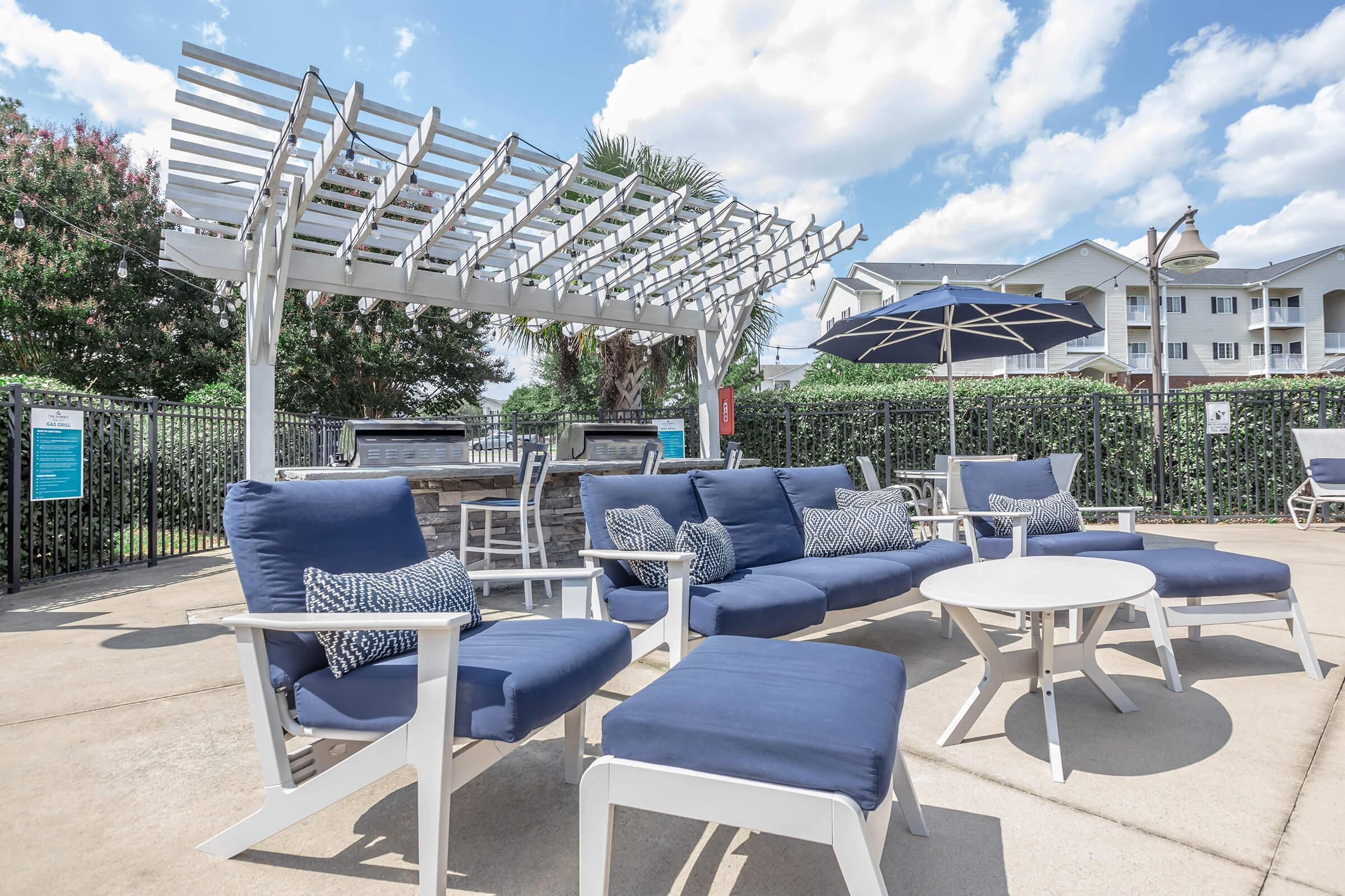 A serene outdoor lounge area featuring blue furniture, including sofas with decorative pillows, a round table, and an umbrella. A pergola provides shade, surrounded by greenery and a pool in the background. Fluffy white clouds are scattered in the blue sky. Ideal for relaxation and social gatherings.