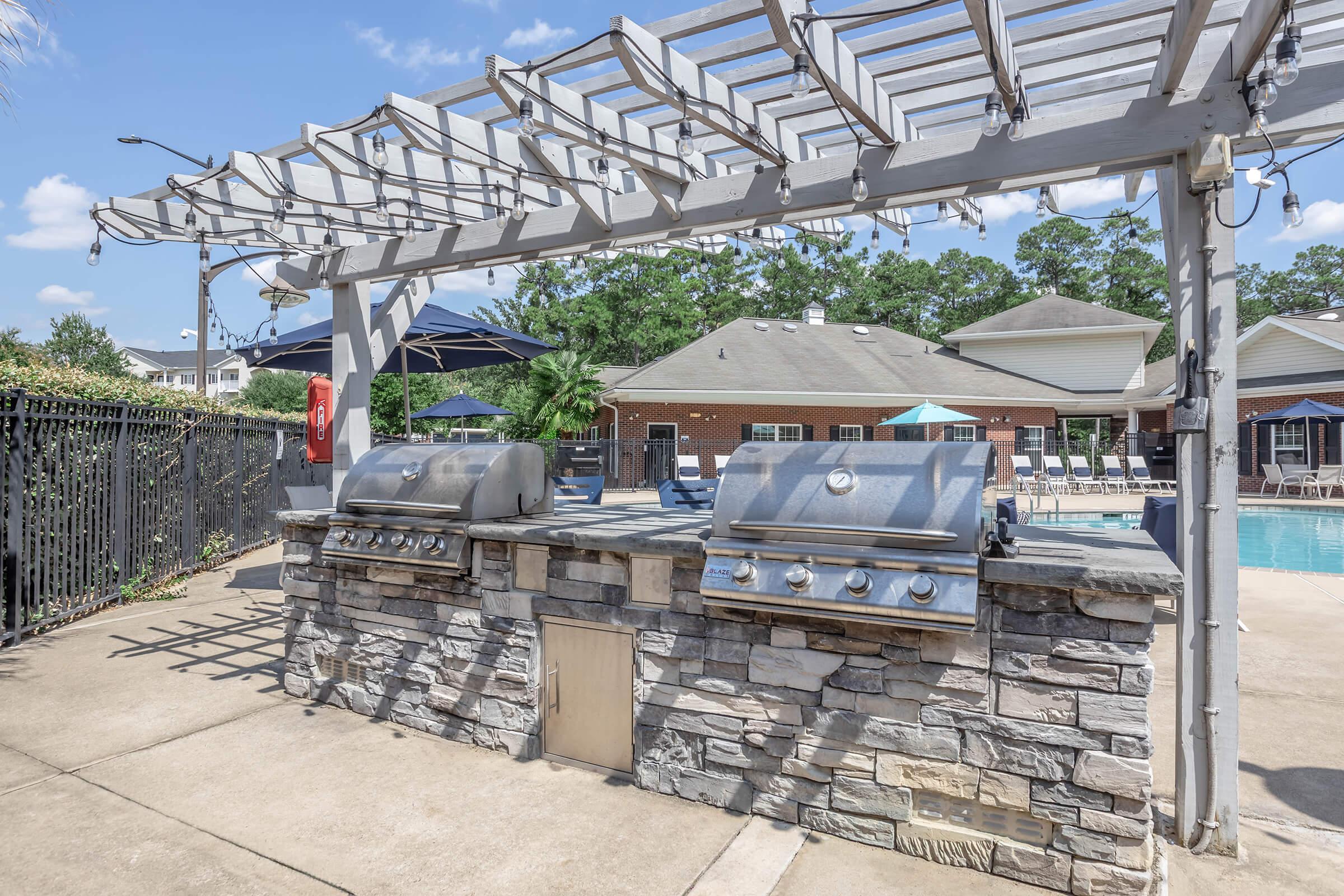 Outdoor grilling area with two stainless steel barbecue grills, situated beside a swimming pool surrounded by lounge chairs and umbrellas. The area features a shaded pergola and brick stonework, creating an inviting space for outdoor cooking and gatherings. Lush greenery is in the background.