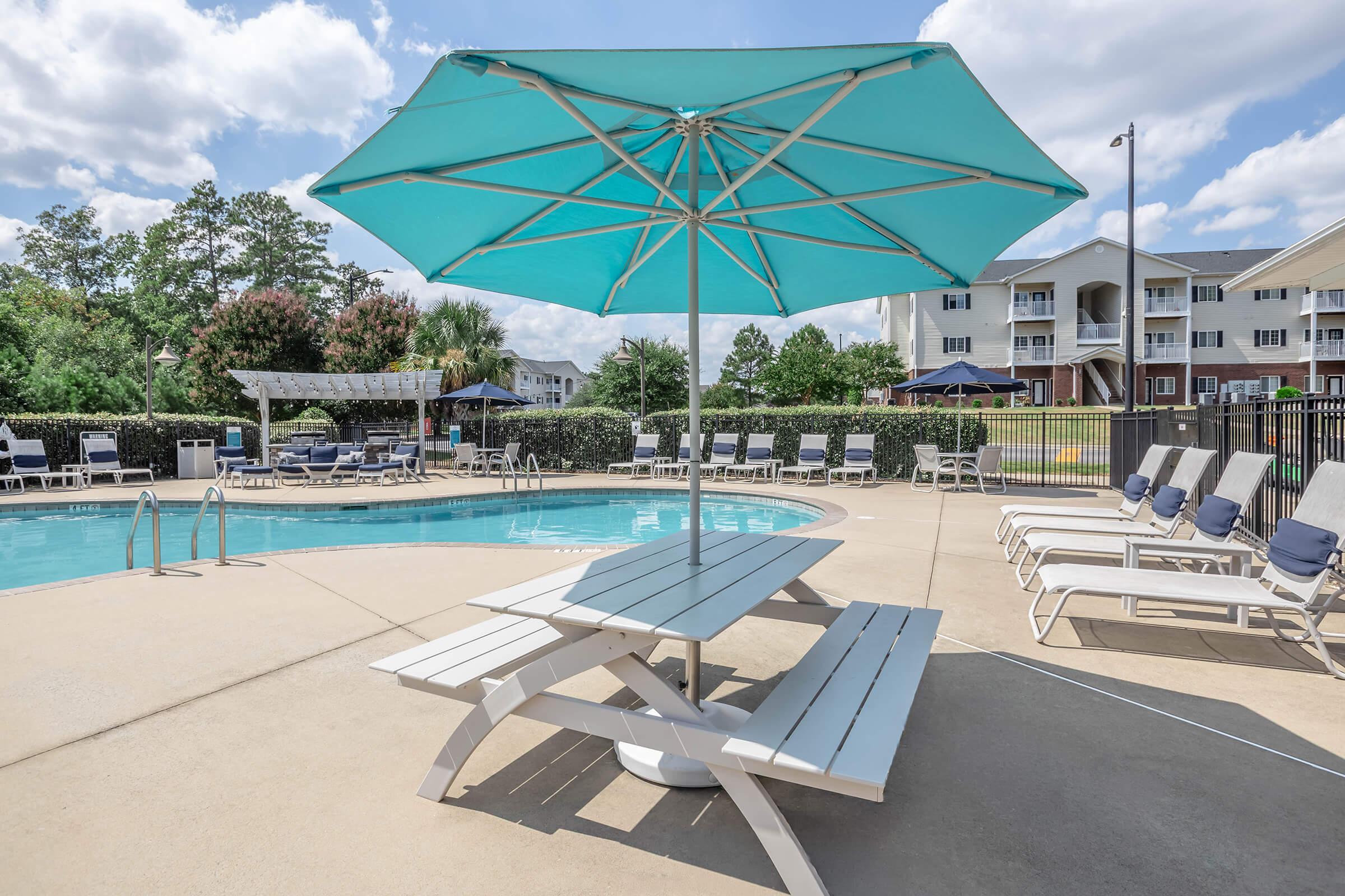 A turquoise umbrella provides shade over a picnic table near a swimming pool. Surrounding the pool are lounge chairs, and greenery is visible in the background, along with buildings. The sky is partly cloudy, creating a bright and inviting atmosphere for outdoor relaxation.