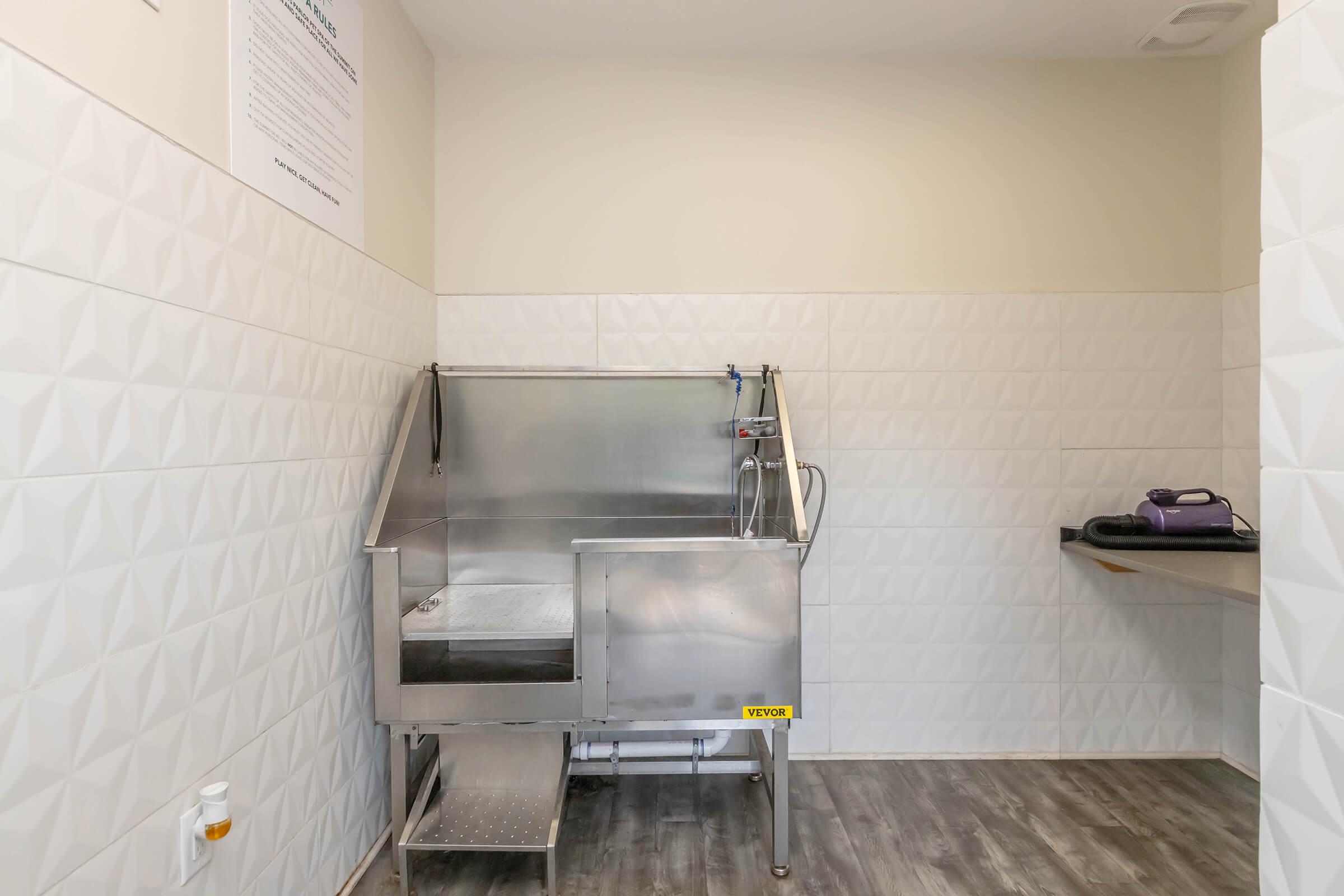 A stainless steel grooming tub with a detachable showerhead, located in a well-lit room with textured white tiled walls. There is a metal step for easy access, and a small countertop on the right side. The space is clean and designed for pet grooming.