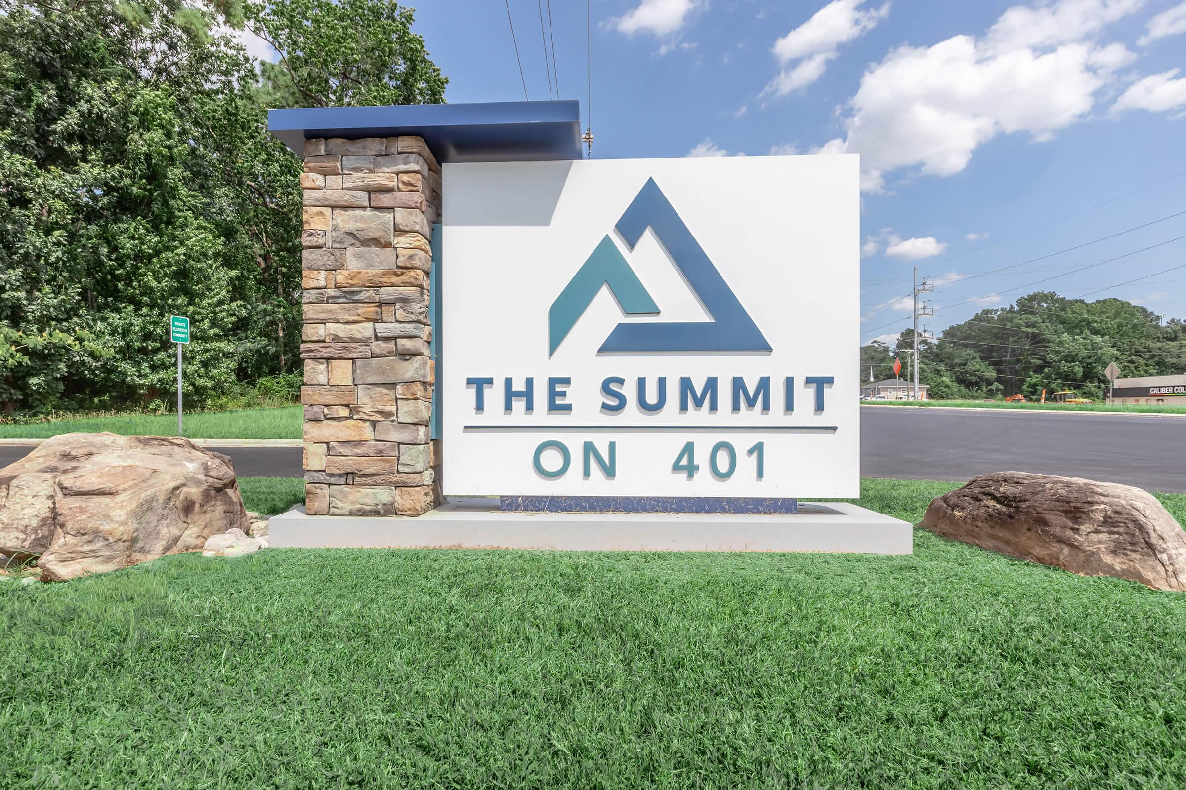Sign for "The Summit on 401" featuring a stylized triangular logo, with a stone and white background. The surrounding area includes green grass and trees, set against a clear blue sky with some clouds.