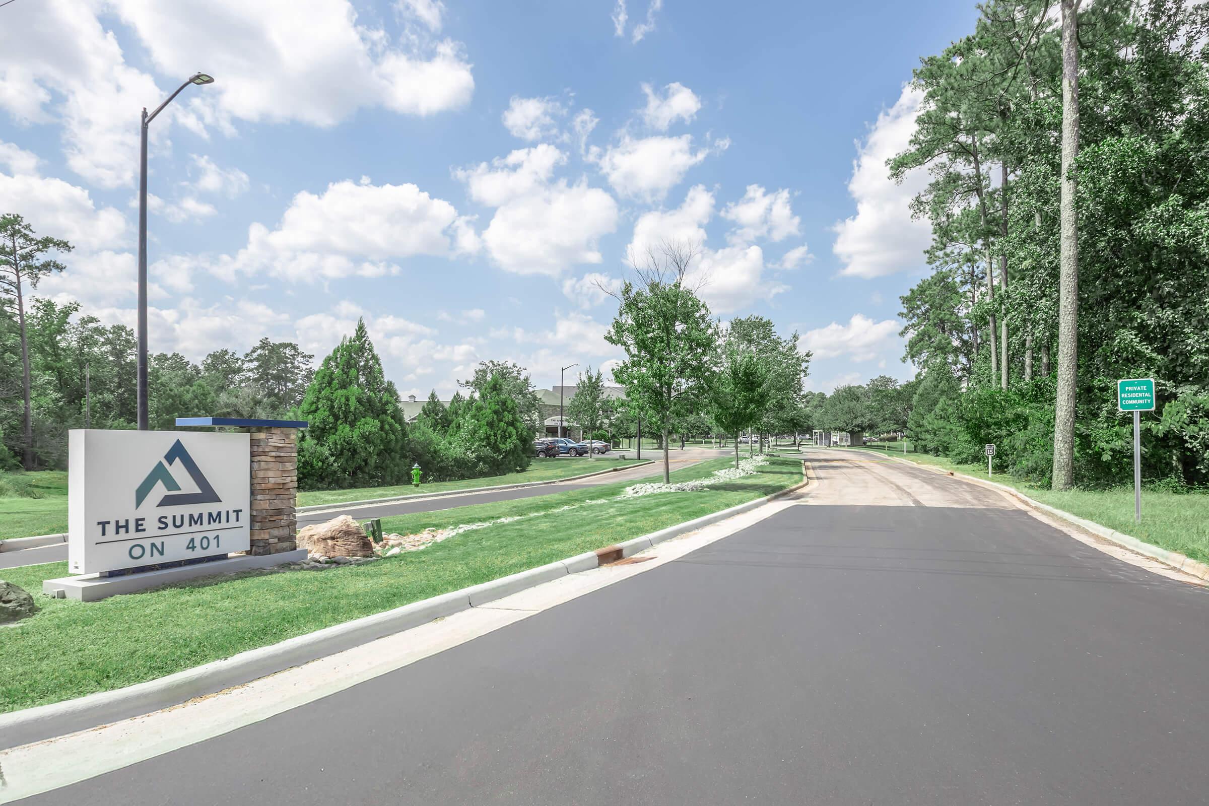 A sunny day scene featuring a paved road leading to a residential area. On the left, a sign reads "The Summit on 401," indicating the location. Lush green grass and trees line the road, with a few cars parked in the background under a blue sky dotted with fluffy white clouds.