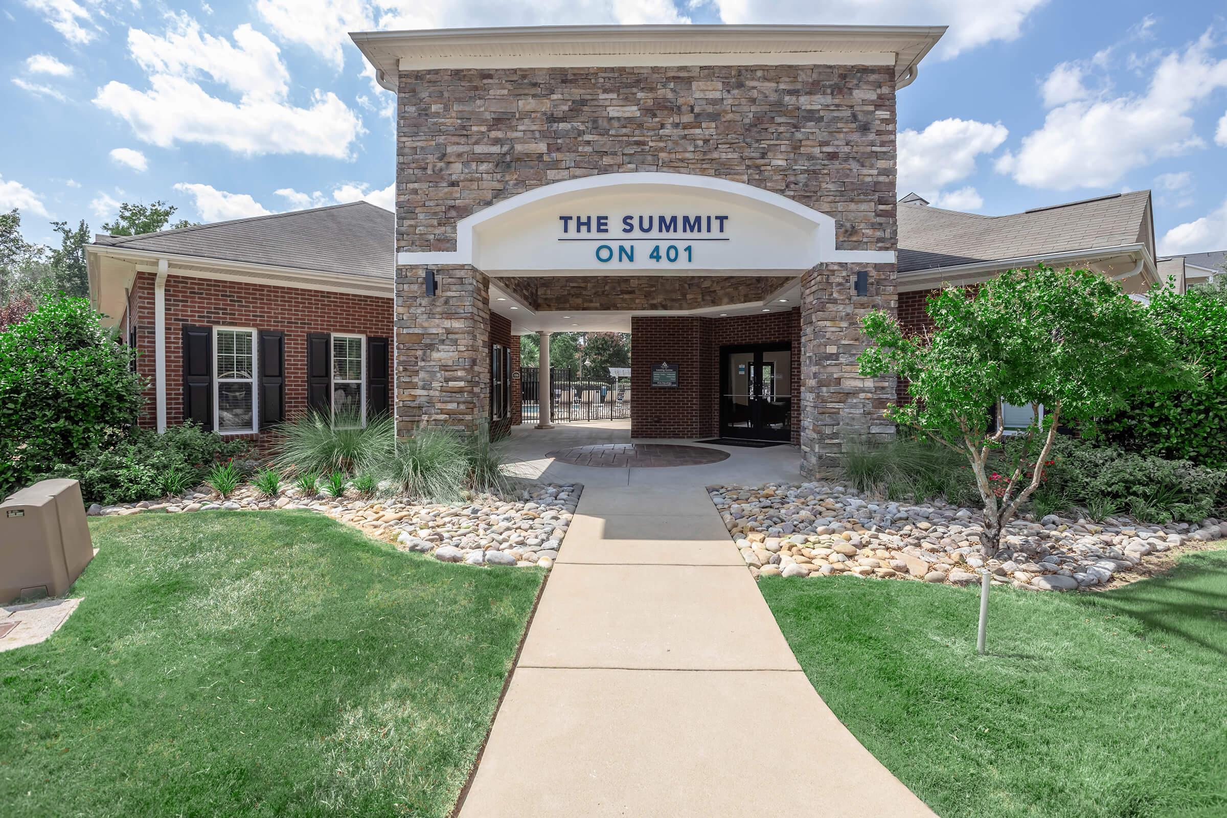 Entrance to "The Summit on 401," featuring a stone and brick façade. A pathway leads to the entrance, bordered by landscaped greenery, shrubs, and decorative stones. The sky is partly cloudy, enhancing the welcoming atmosphere of the building.