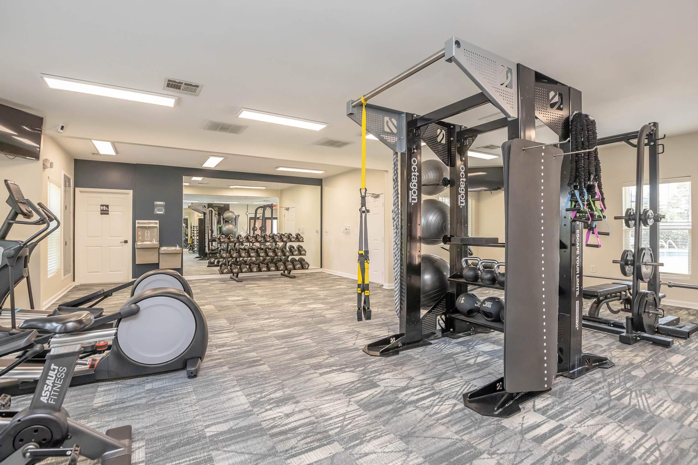 A modern gym interior featuring exercise equipment including an elliptical machine, weight machines, and a functional training rig with suspension straps and weights. The space is well-lit with large mirrors and a clean, open layout, designed for fitness activities and workouts.