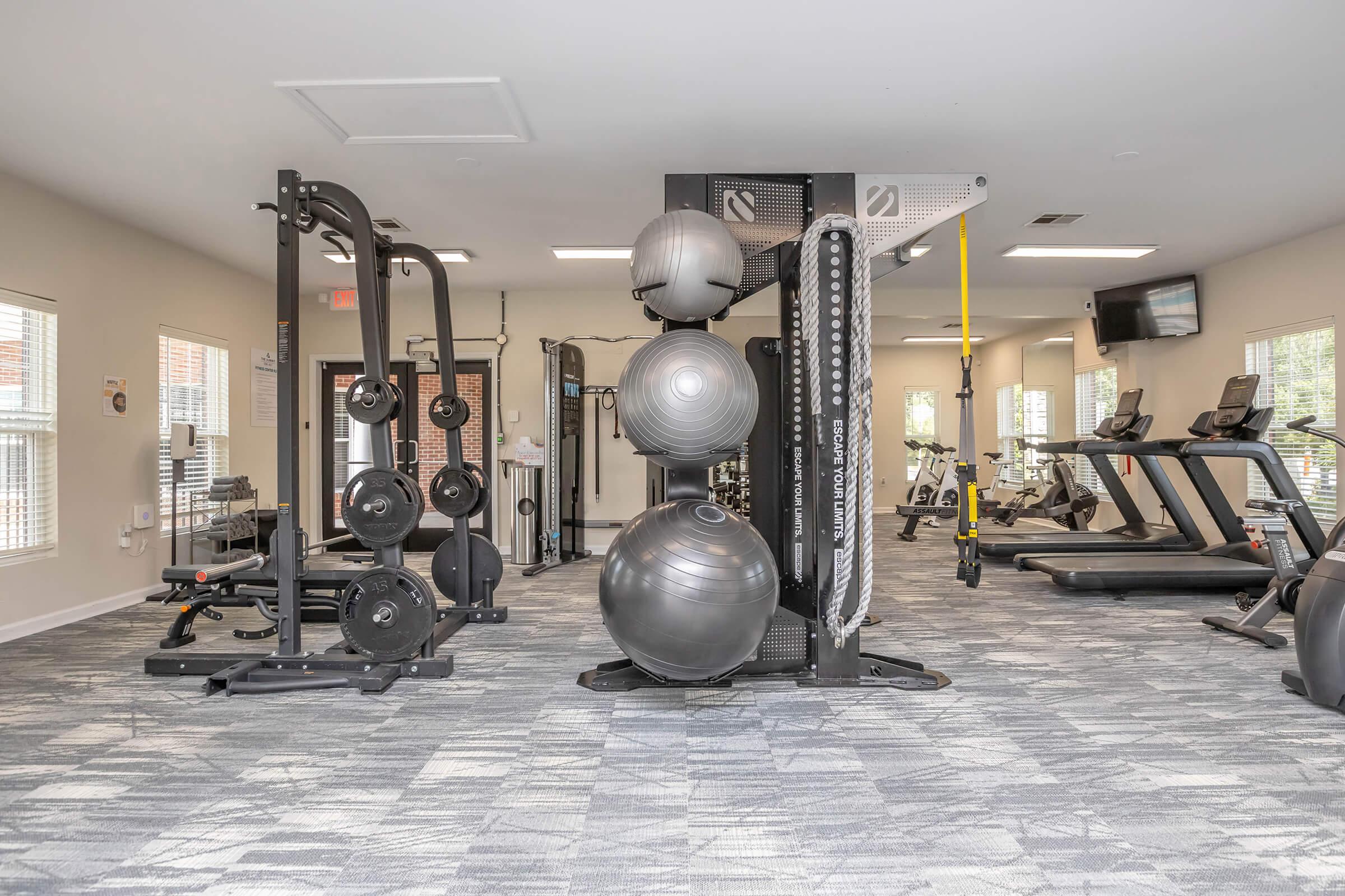 A modern gym interior featuring various exercise equipment, including weight machines, free weights, kettlebells, stability balls, and cardio machines like treadmills. The space is well-lit with large windows and has a neutral color scheme and carpeted flooring, creating a welcoming atmosphere for workouts.