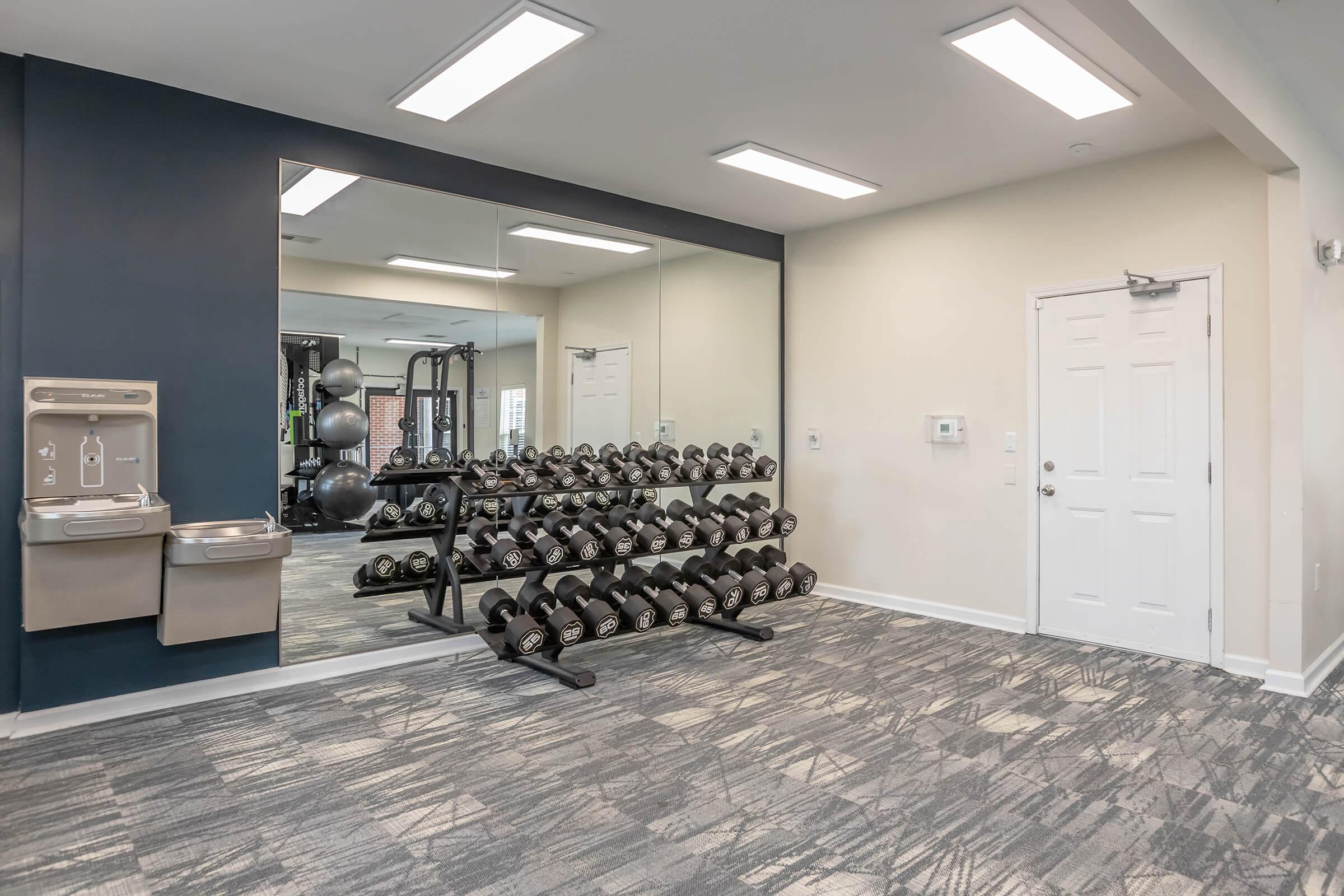 Interior of a modern gym featuring a rack of free weights, a water fountain, and a door. The walls are painted in neutral tones with a large mirror reflecting workout equipment in the background. The flooring is a textured carpet, and the space is well-lit with overhead lights.