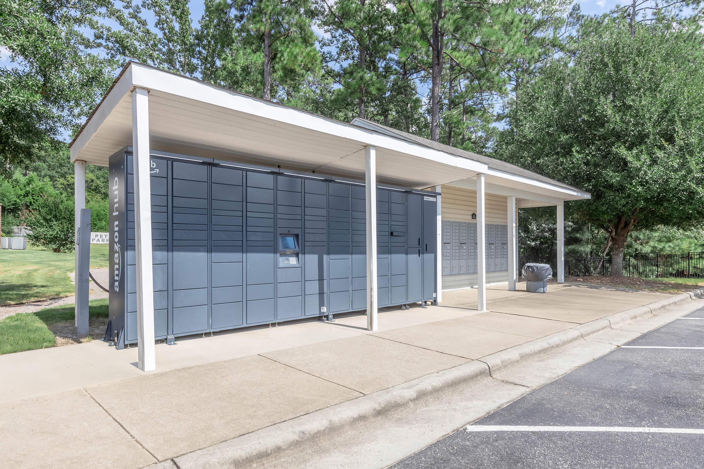 A row of modern parcel lockers under a covered shelter, surrounded by greenery. The structure features a large gray automated locker unit with a touch screen, alongside traditional mailboxes. The driveway is paved, and the scene is sunny with clear skies.