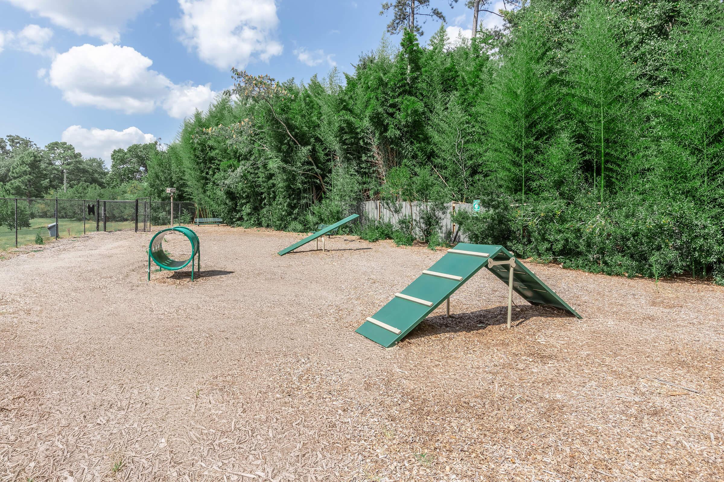 A dog agility training area featuring a climbing ramp, a tunnel, and a seesaw, set in a spacious, gravel-filled outdoor environment surrounded by green trees and shrubs under a partly cloudy sky.