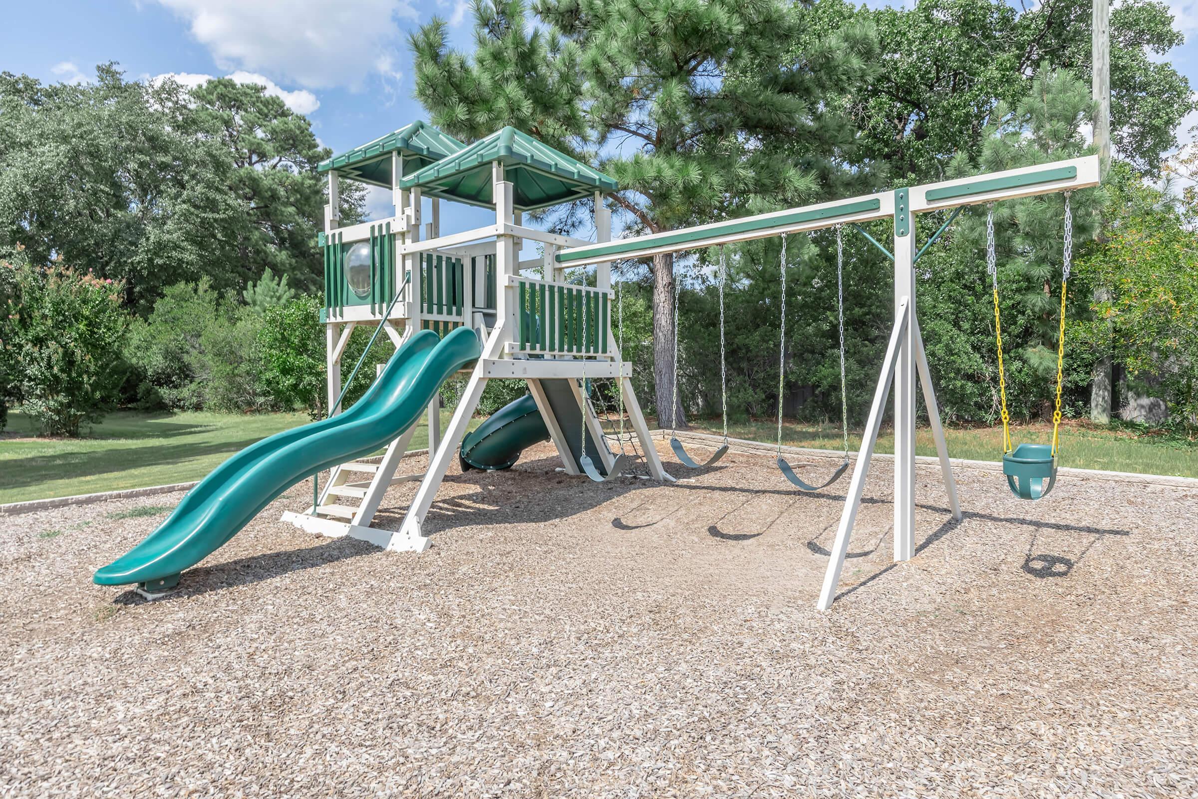 A children's playground featuring a green slide, a multi-level play structure with a roof, swings, and a baby swing, surrounded by a grassy area and trees. The ground is covered with wood chips, providing a safe play surface.