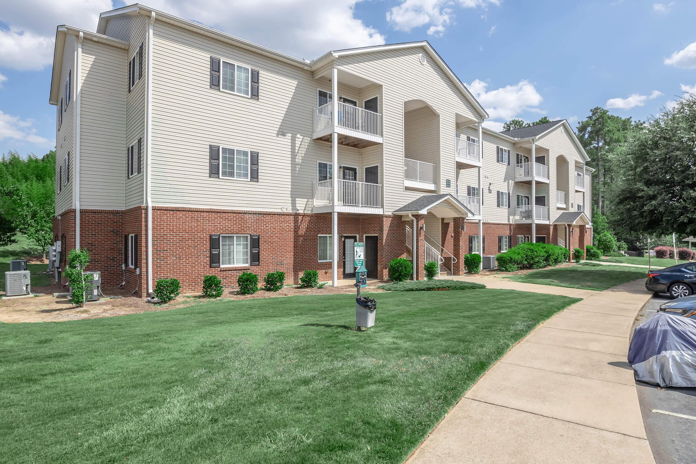 A three-story apartment building with beige siding and red brick accents, surrounded by manicured lawns and small shrubs. The building features several balconies, and a walkway leads to the entrance. Clear blue skies are visible in the background.