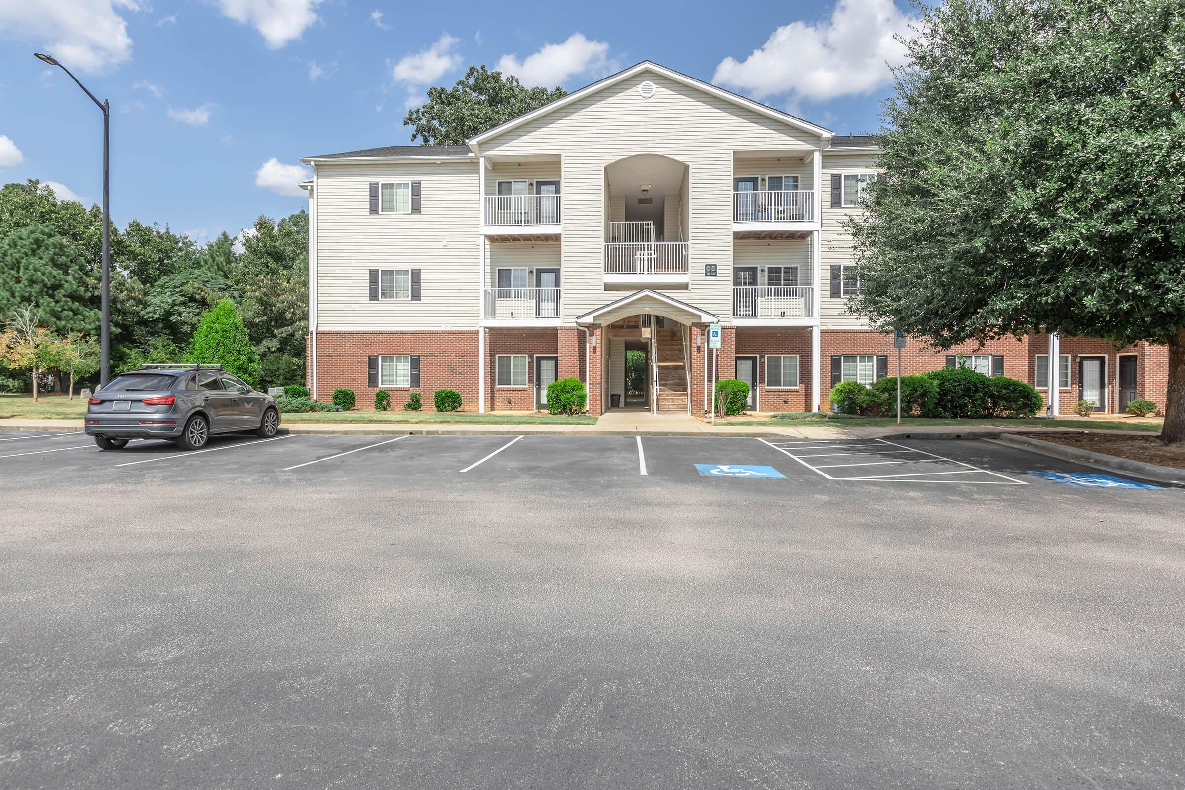 A multi-story residential building with a beige exterior and brick accents. The building has a central entrance, surrounded by trees and landscaped shrubs. In the foreground, there is a parking lot with marked parking spaces, including a designated handicap spot. The sky is partly cloudy.