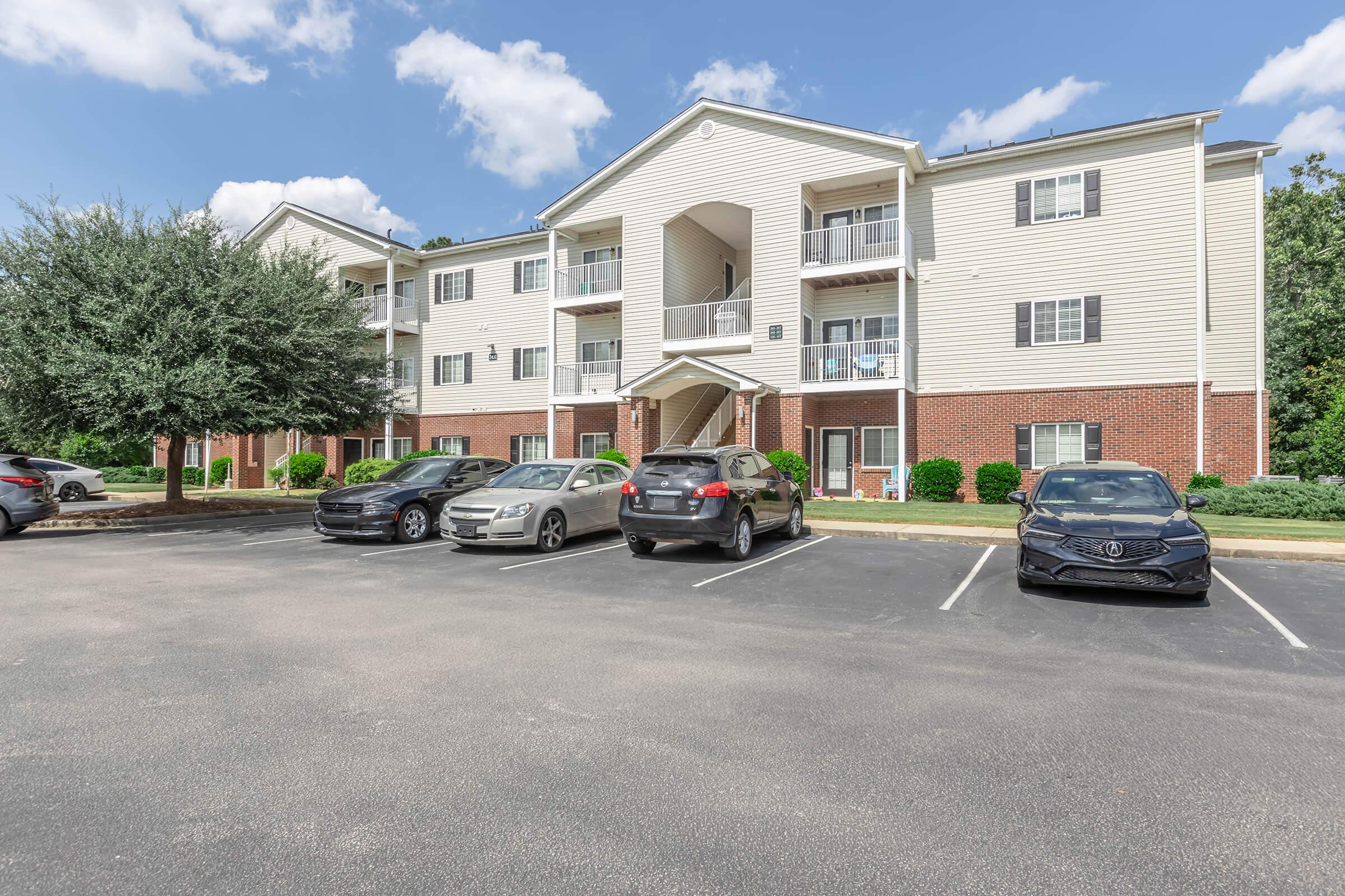 A three-story apartment building with a mix of brick and siding exterior. The building features multiple balconies and surrounded by greenery. Several cars are parked in a lot in front of the building, under a partly cloudy sky.