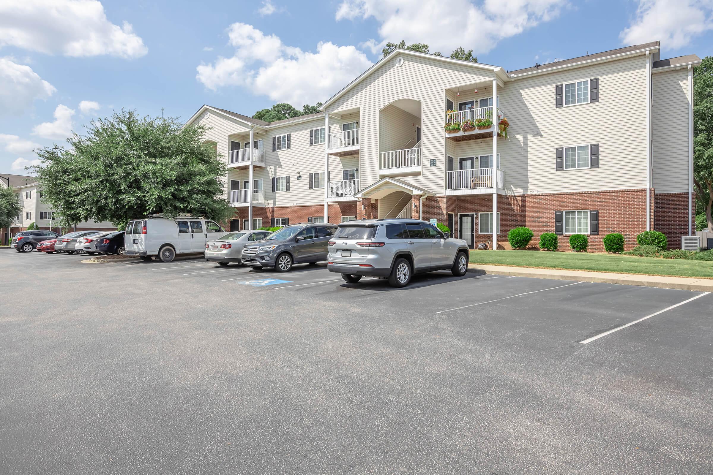A residential apartment building with multiple levels, featuring balconies with potted plants. Several parked cars are visible in the foreground on a paved lot, with trees and shrubs adding greenery around the building. The sky is partly cloudy, creating a bright and inviting atmosphere.