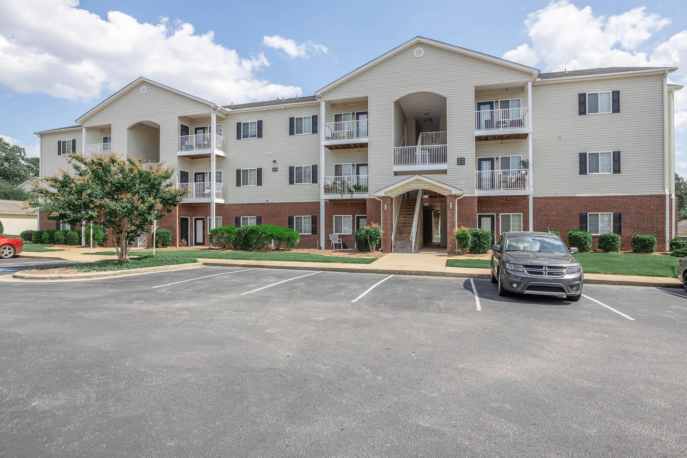 A three-story residential apartment building with a beige exterior and brick accents. It features multiple balconies, green shrubs, and a parking area with several cars. The sky is partly cloudy, creating a bright atmosphere.
