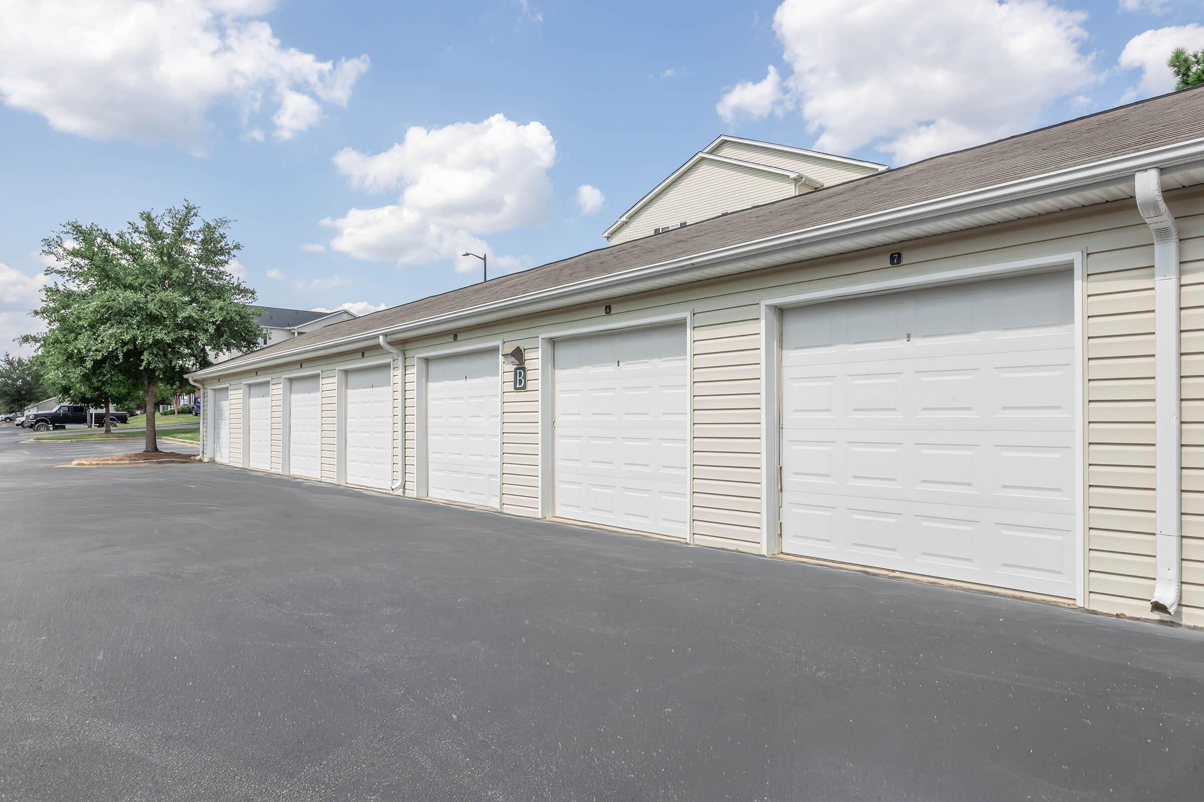 A row of white garage doors on a light-colored building, set against a clear blue sky with fluffy white clouds. The pavement is black, and there are trees in the background, providing a suburban or residential feel. The garages are neatly aligned, with some numbers visible on the doors.