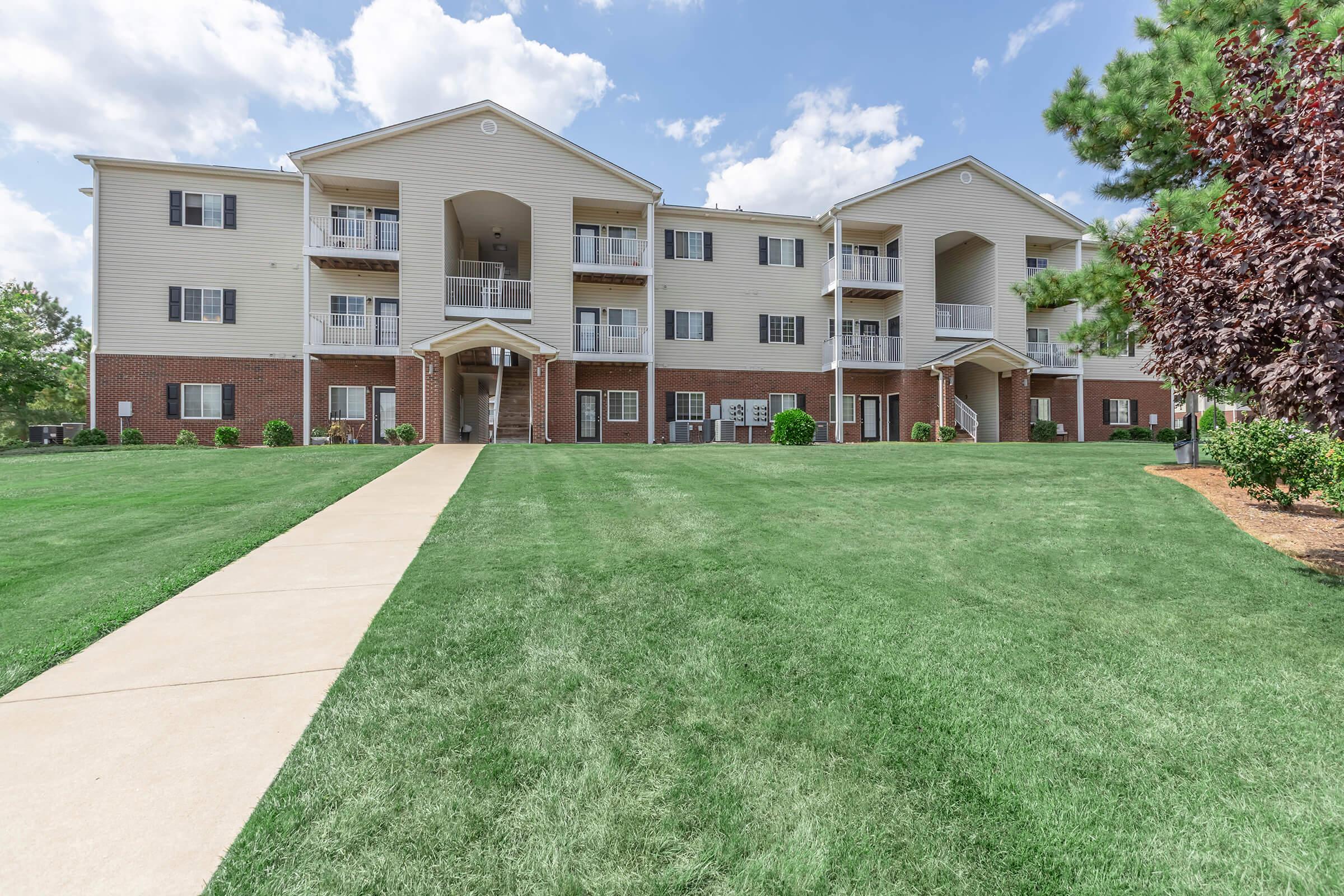 A three-story residential building with a light-colored exterior, featuring multiple balconies and large windows. The well-maintained lawn in front is lush and green, lined with small bushes. The sky is partly cloudy, creating a pleasant atmosphere. A paved walkway leads to the main entrance.