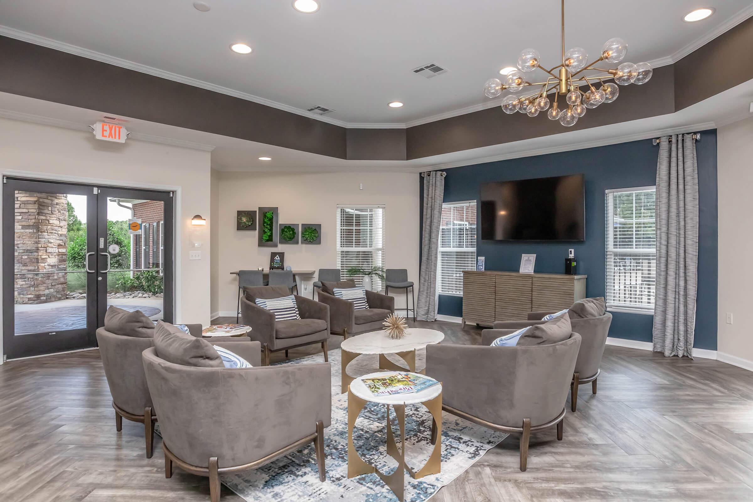 A modern living area featuring comfortable gray armchairs arranged around a light-colored coffee table. A stylish chandelier hangs from the ceiling. There’s a large wall-mounted TV and decorative plants on the wall. Large windows allow natural light in, and an entrance leads outside.