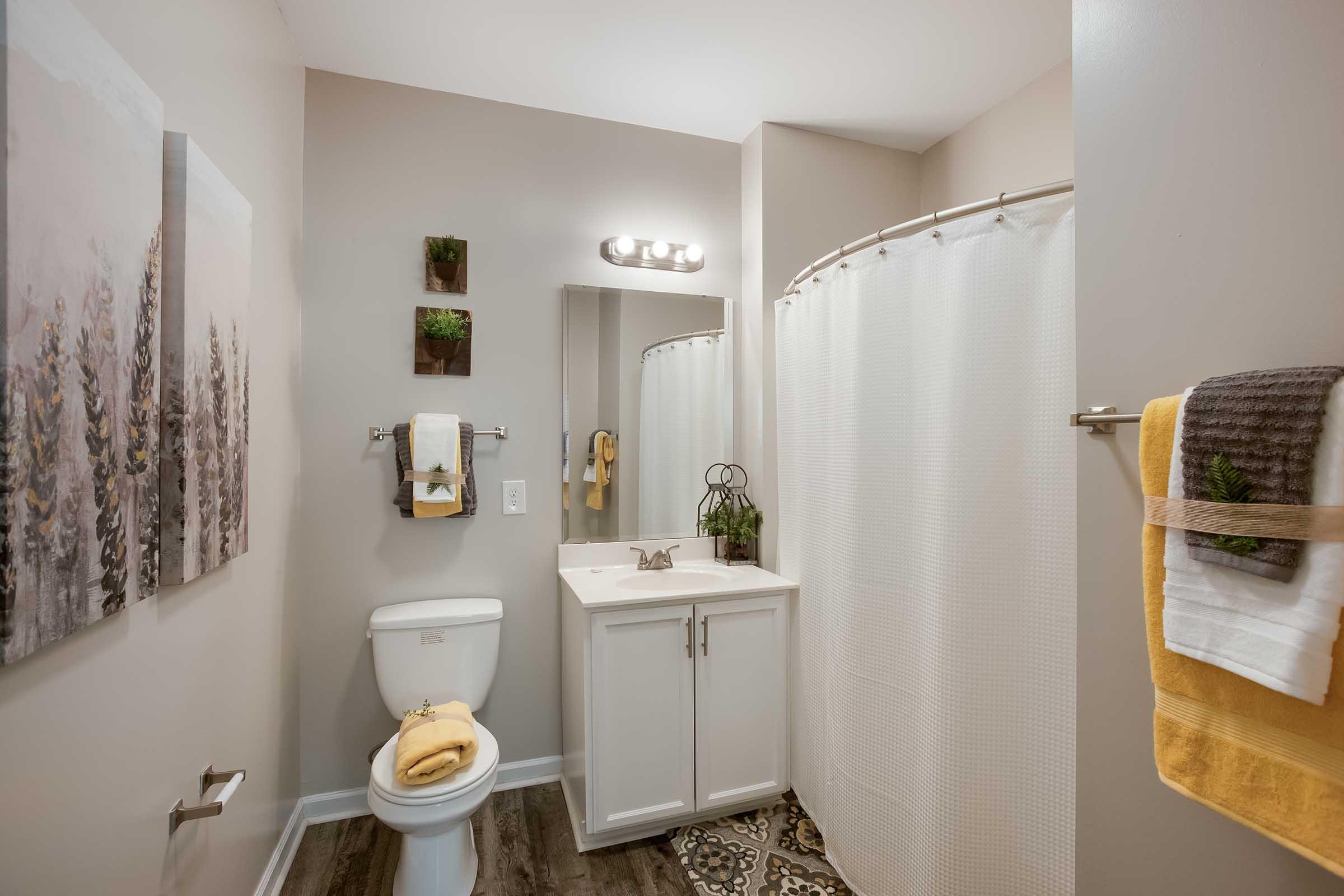 A modern bathroom featuring a white toilet, a sink with a mirror above, and a shower with a white curtain. Decorated with wall art of trees, small potted plants, and neatly arranged towels in yellow and gray. The floor has a patterned design and the walls are painted light beige.