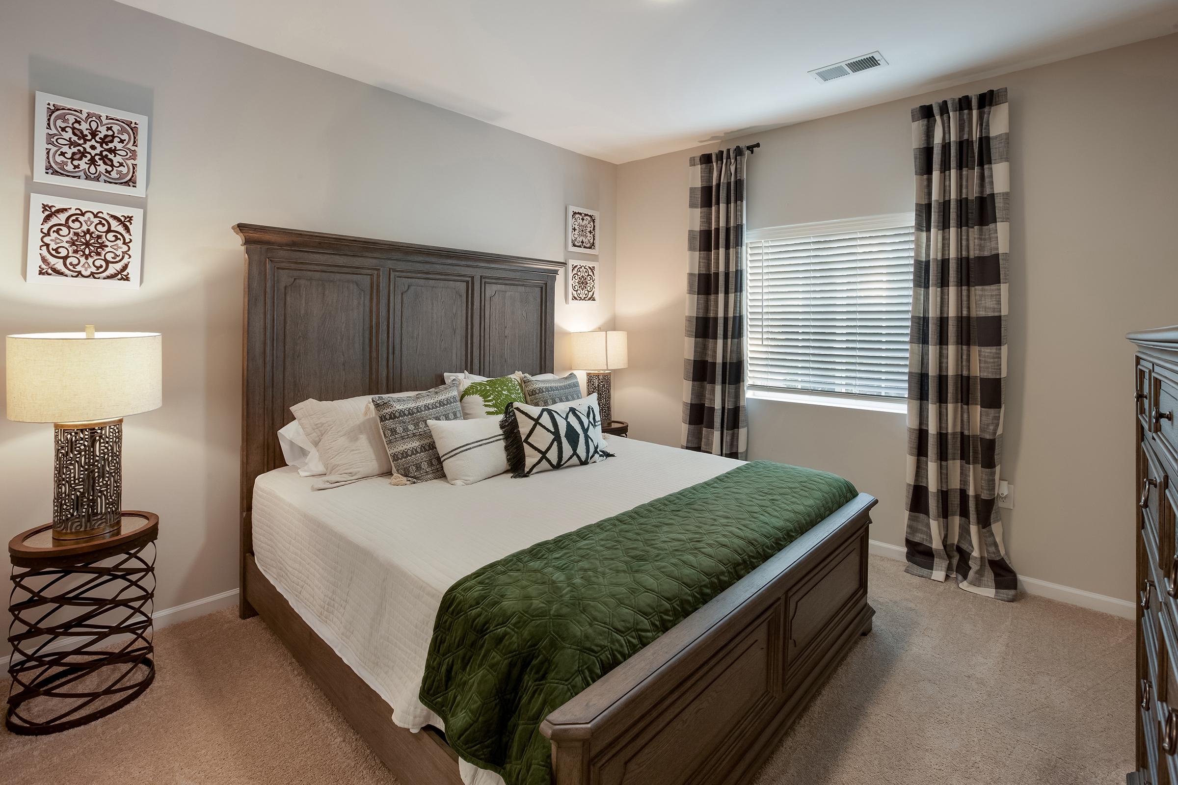A cozy bedroom featuring a large wooden bed with a green throw blanket, decorative pillows, and a stylish headboard. There are two lamps on bedside tables, patterned curtains, and wall art above the bed, along with a window allowing natural light to brighten the space. The carpet adds warmth to the room.