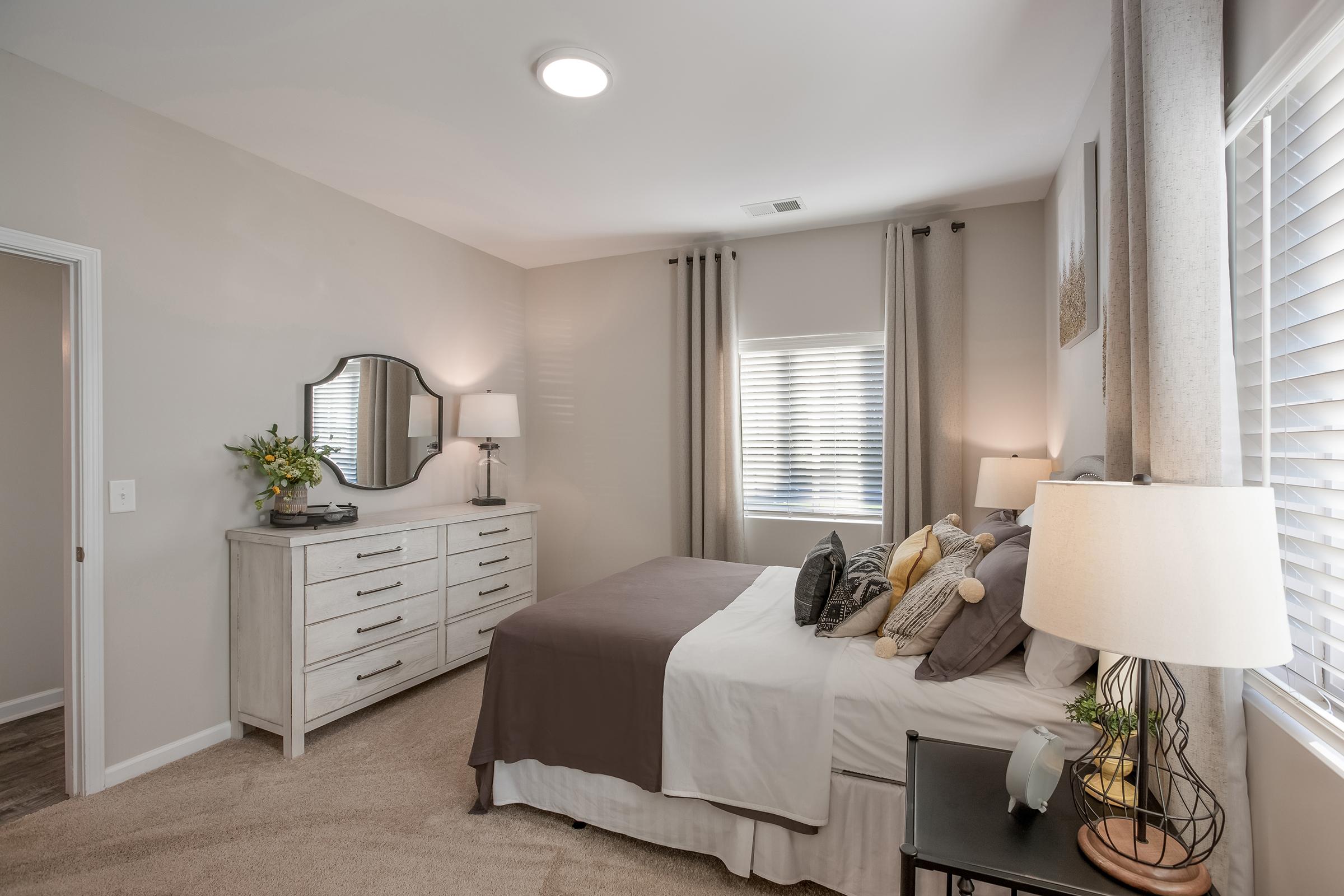 A well-decorated bedroom featuring a queen-sized bed with gray and white bedding. There's a light wooden dresser with a round mirror above it, a bedside table with a lamp, and large windows adorned with light curtains. The walls are painted in soft neutral tones, creating a cozy atmosphere.