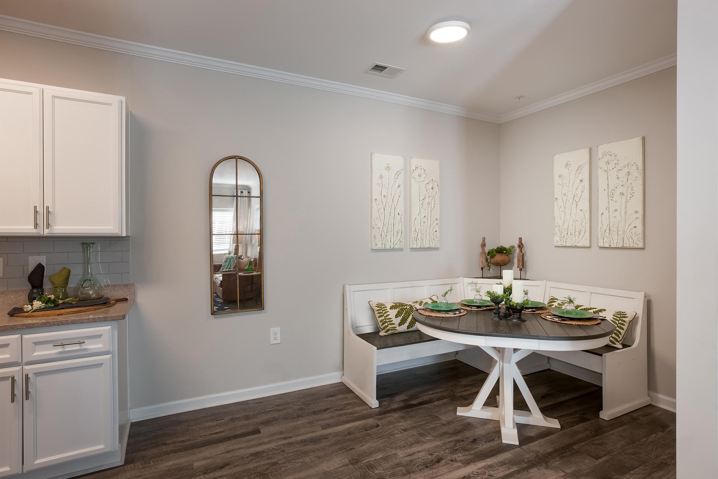 A cozy dining area featuring a round table with decorative plates and green accents. There is a built-in bench with cushions, a mirror on the wall, and two framed wall art pieces. The kitchen area is visible with white cabinetry and a wooden countertop, creating a warm and inviting atmosphere.